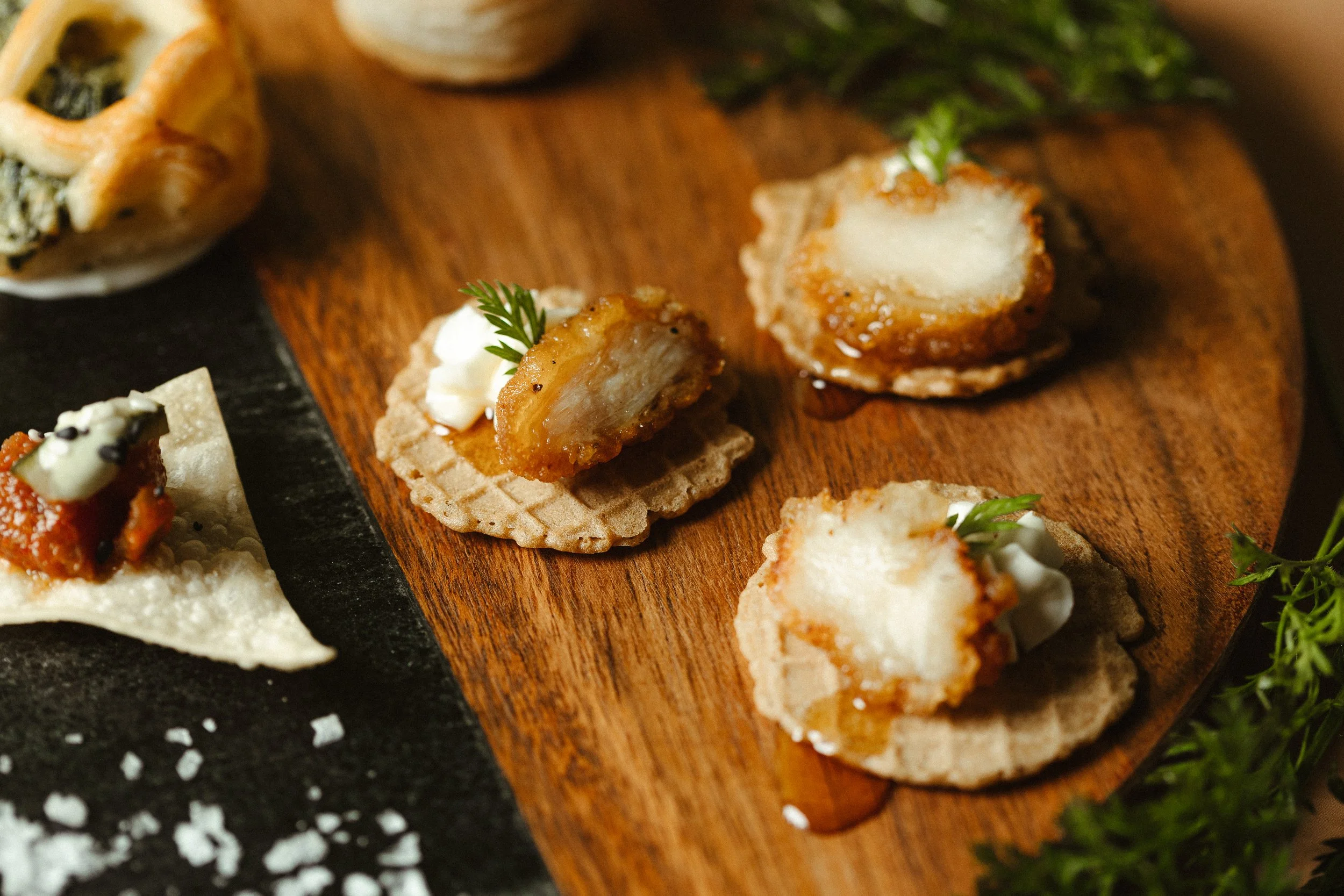 Various small appetizers served on a wooden and slate platter, including fried bites with creamy toppings, garnished with herbs, alongside small bread slices and garnishes.
