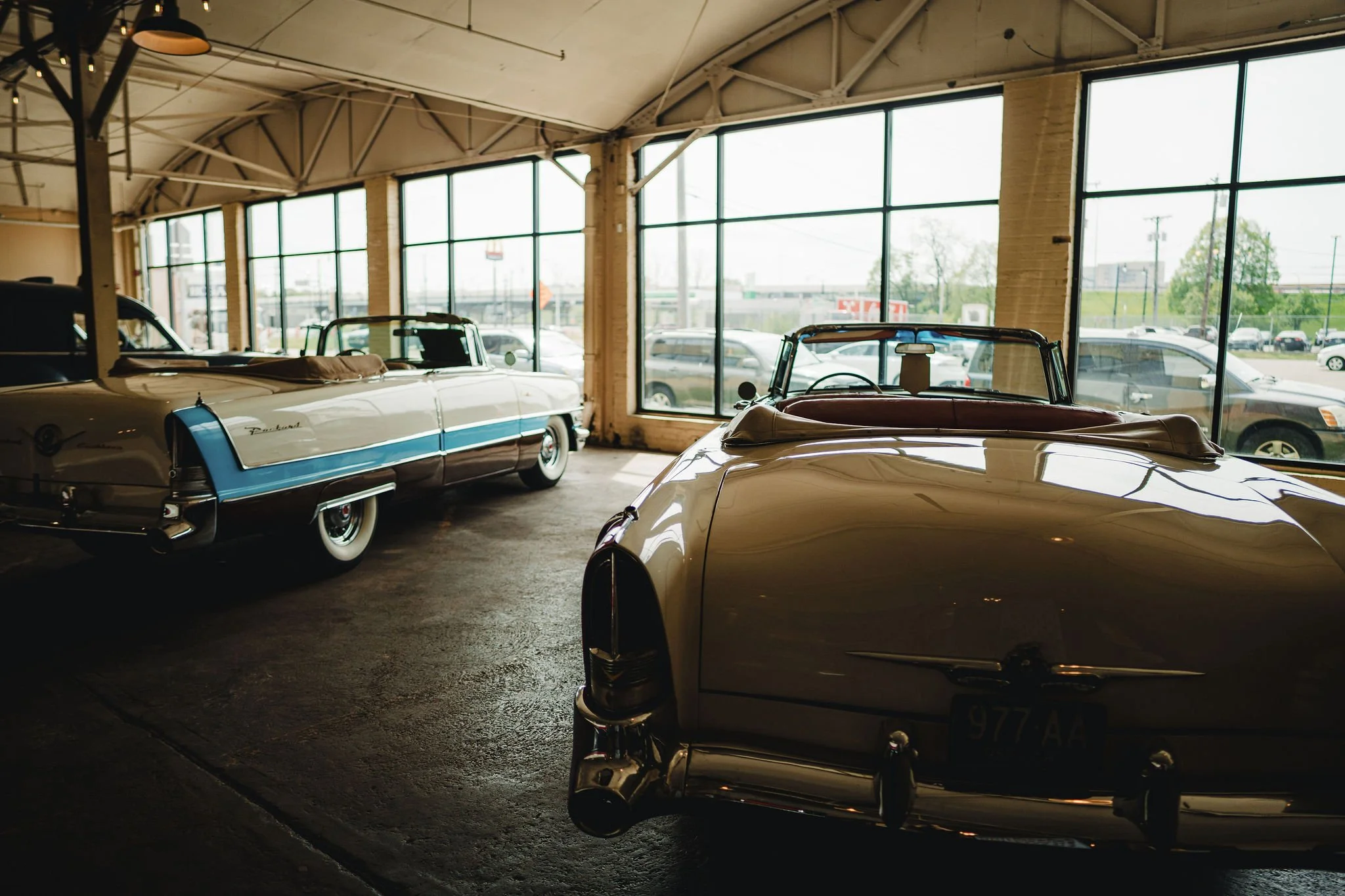 Interior of a vintage car dealership featuring classic cars parked inside near large window panels, with natural light coming through.