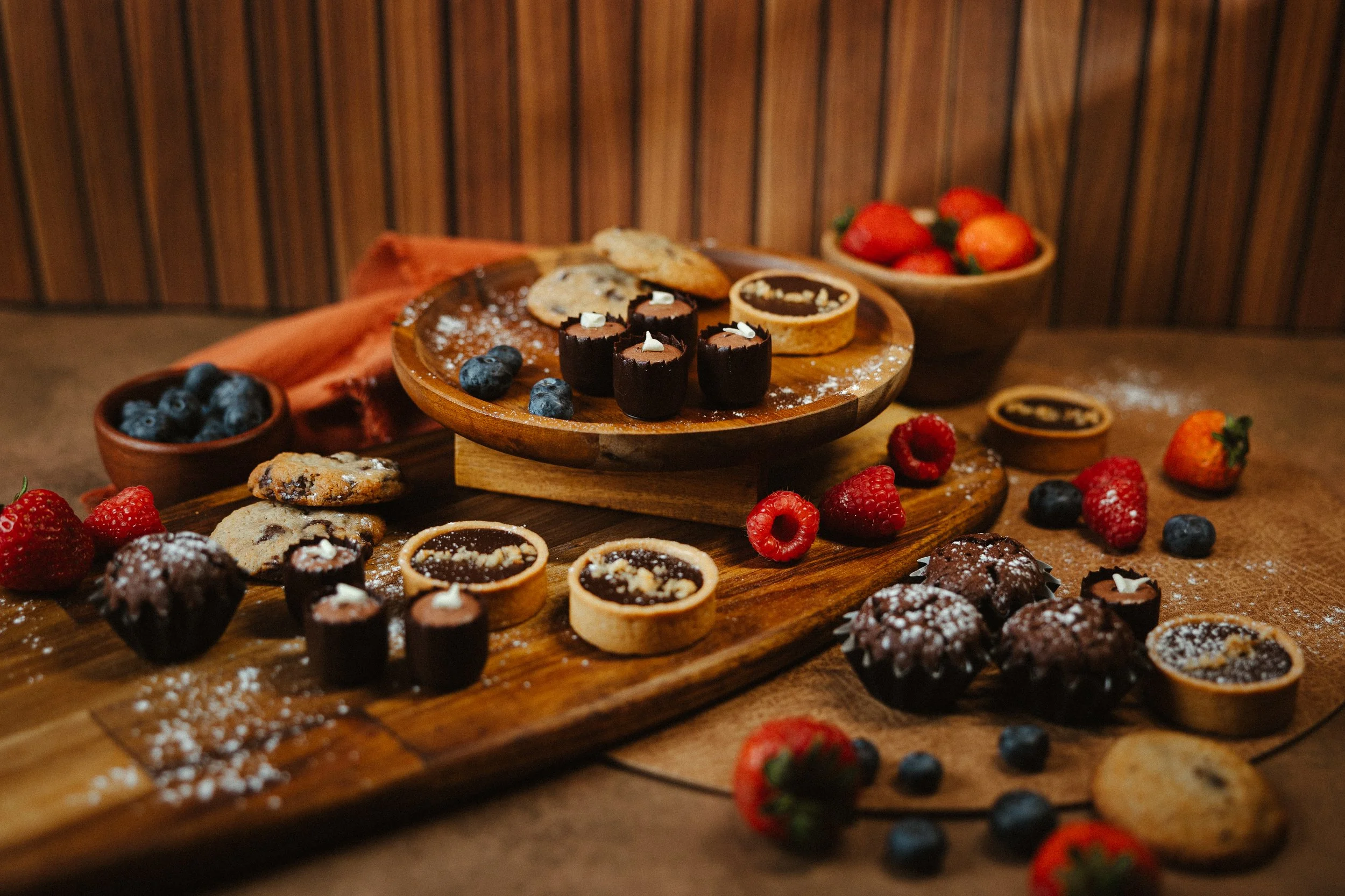 A wooden table with various cookies, berries, and small desserts, including chocolate-filled cookies, strawberries, blueberries, and chocolate tartlets, with a wooden backdrop.