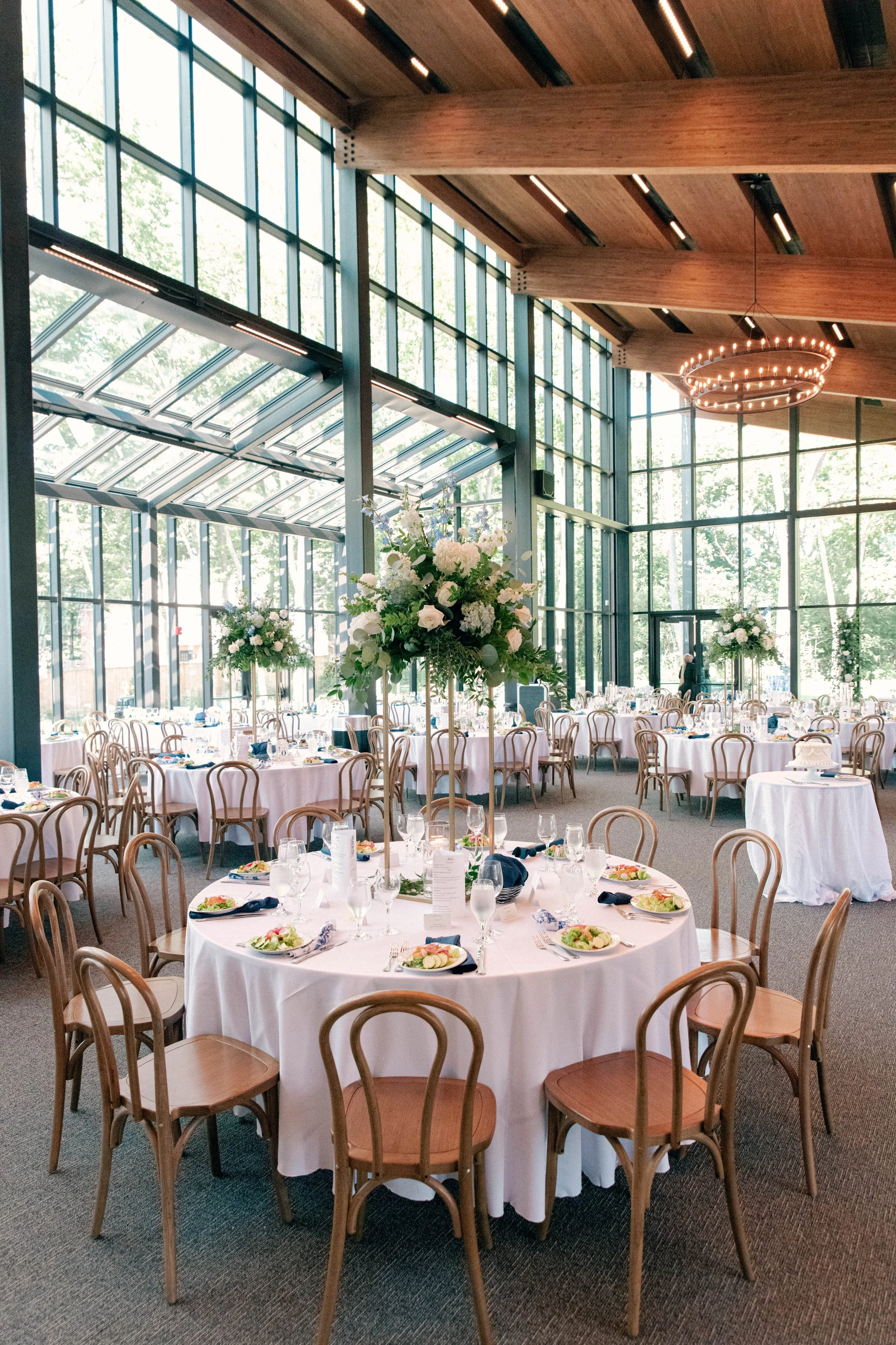 Elegant banquet hall with large glass windows, decorated for a formal event with round tables, white tablecloths, floral centerpieces, and place settings. Wooden ceiling with a chandelier.