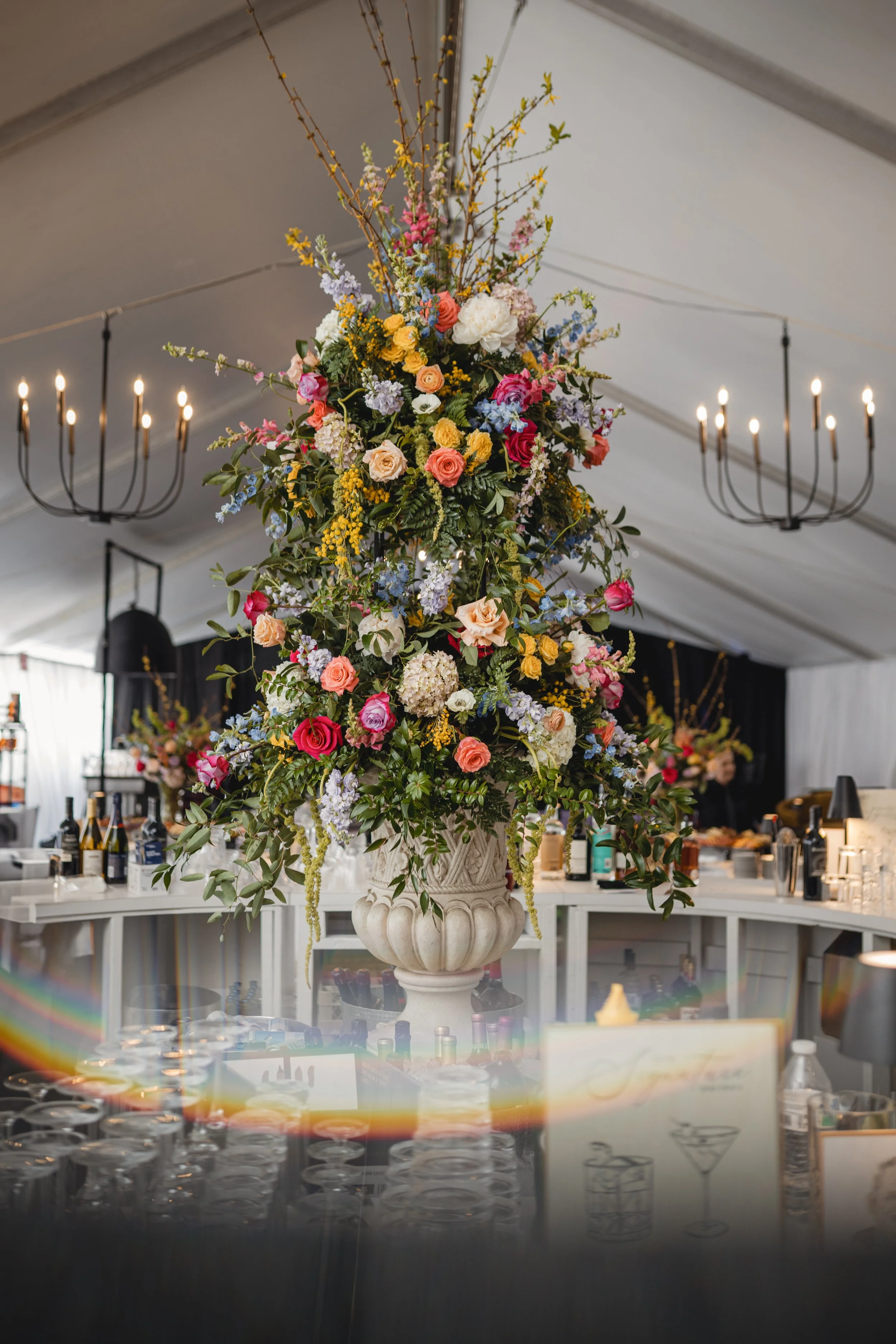 A large floral arrangement with pink, yellow, purple, white flowers and green leaves in a decorative white stone vase, with chandeliers hanging from the ceiling and a bar area with bottles visible in the background.