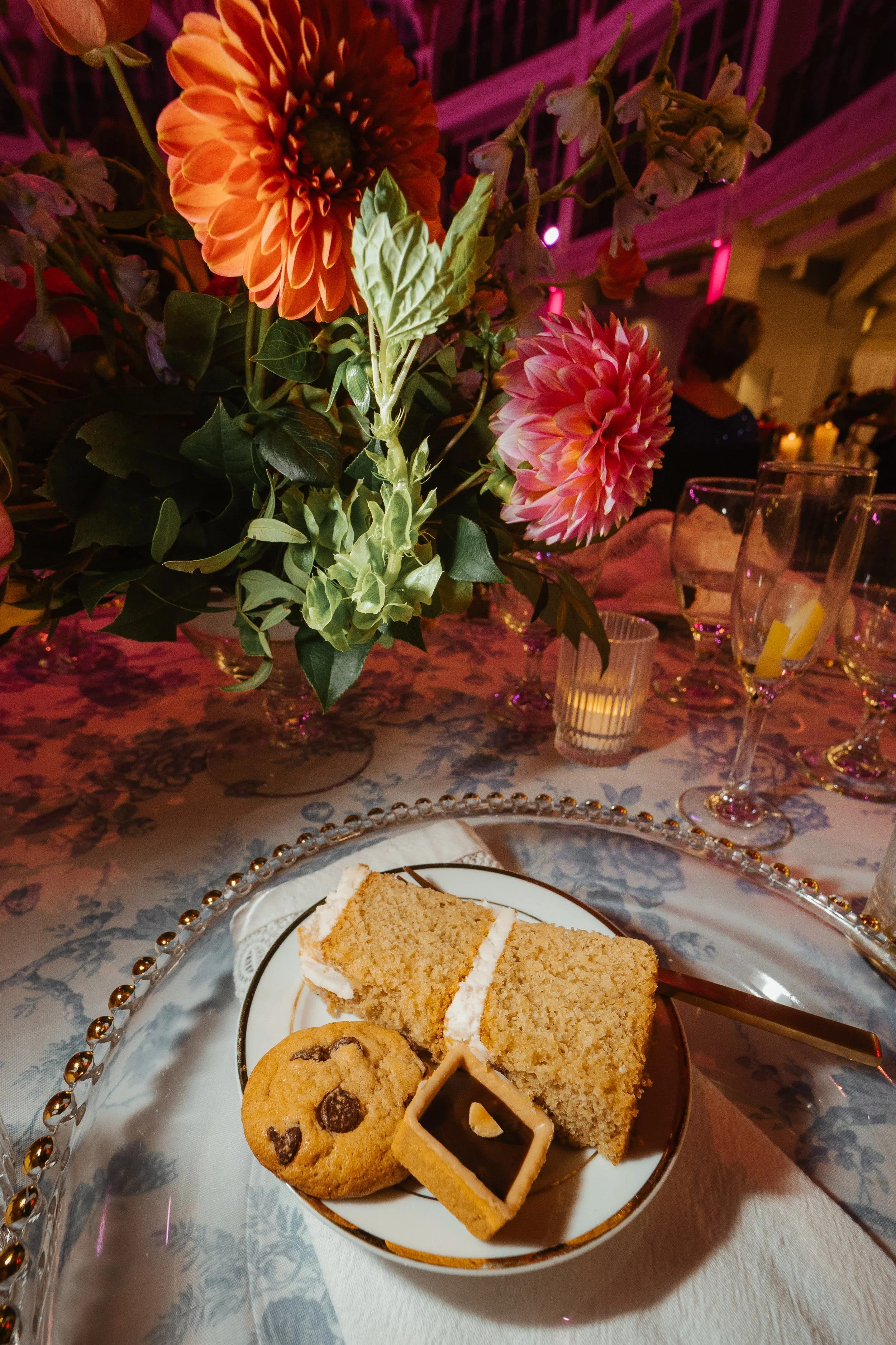 A plate with a slice of cake, cookies, and a decorated cookie on a fancy glass plate, with a floral centerpiece featuring orange and pink flowers, on a table with a patterned tablecloth at an indoor event.