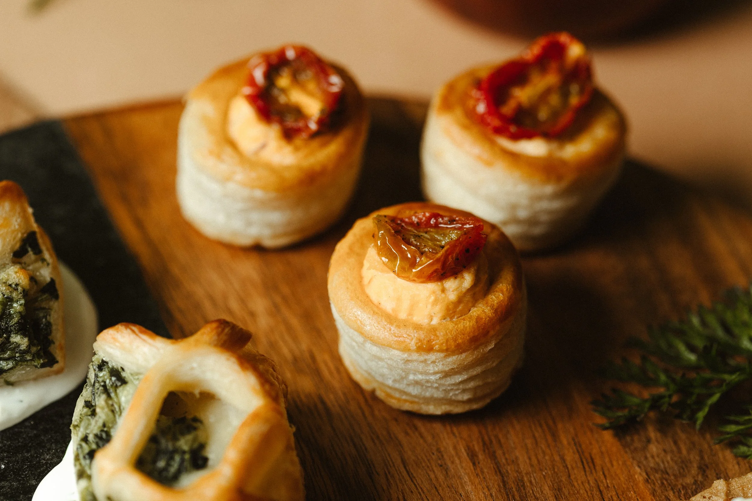 Close-up of assorted appetizers on a wooden platter, including bite-sized rolls topped with sun-dried tomatoes and cheese, and other various snacks.