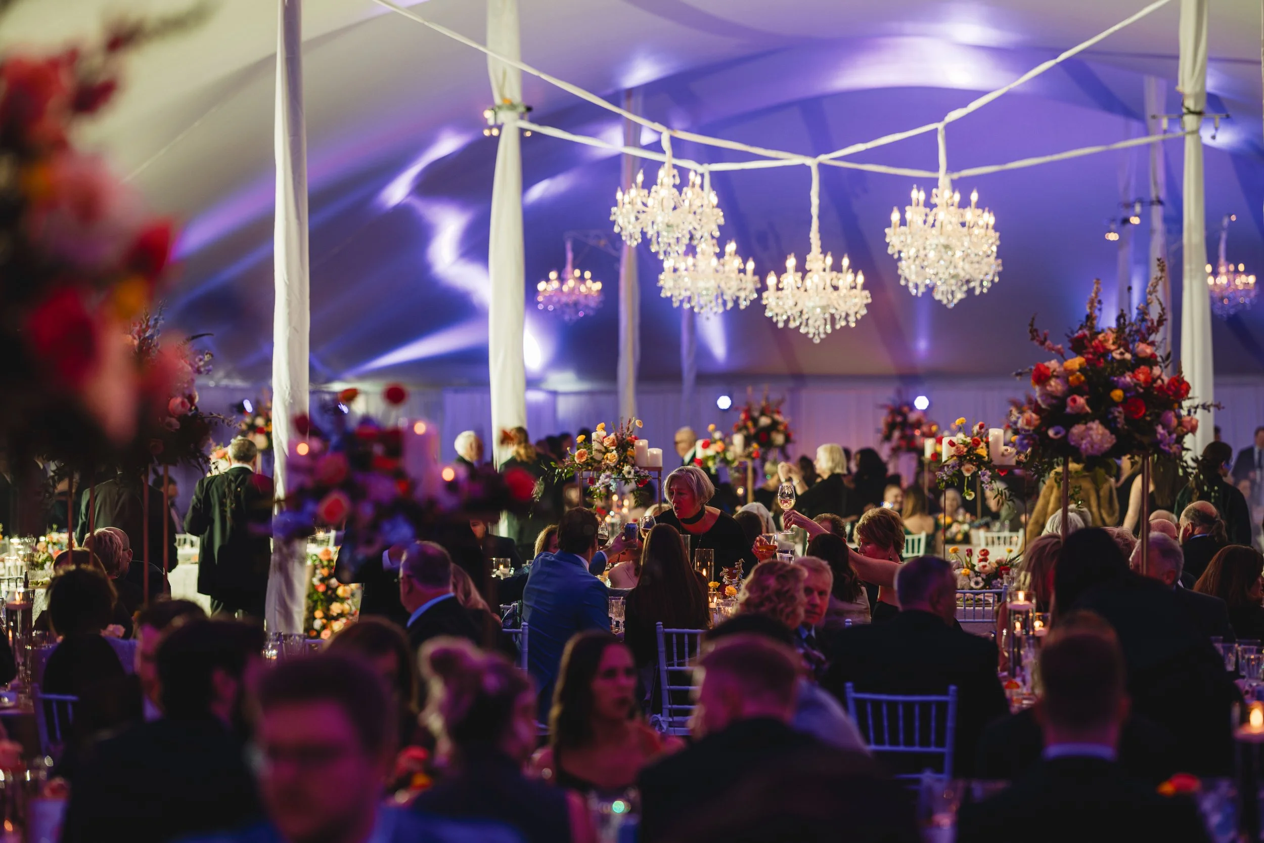 A large indoor event with chandeliers hanging from a decorated ceiling, surrounded by purple and pink lighting. Guests are seated at tables with floral centerpieces, and some people are standing or serving drinks. The atmosphere appears festive and elegant.