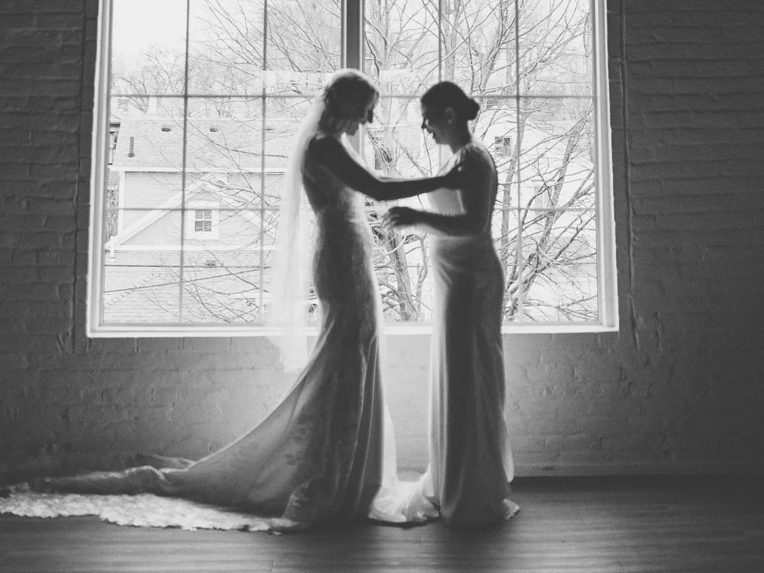 Two women in wedding dresses standing close by a large window, holding hands, with a wintery scene of leafless trees and rooftops outside.