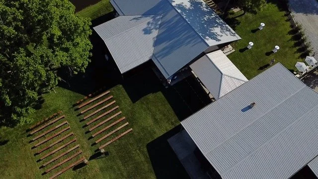 Aerial view of outdoor space, a large tree, and buildings with metal roofs, along with white outdoor furniture and a nearby street.