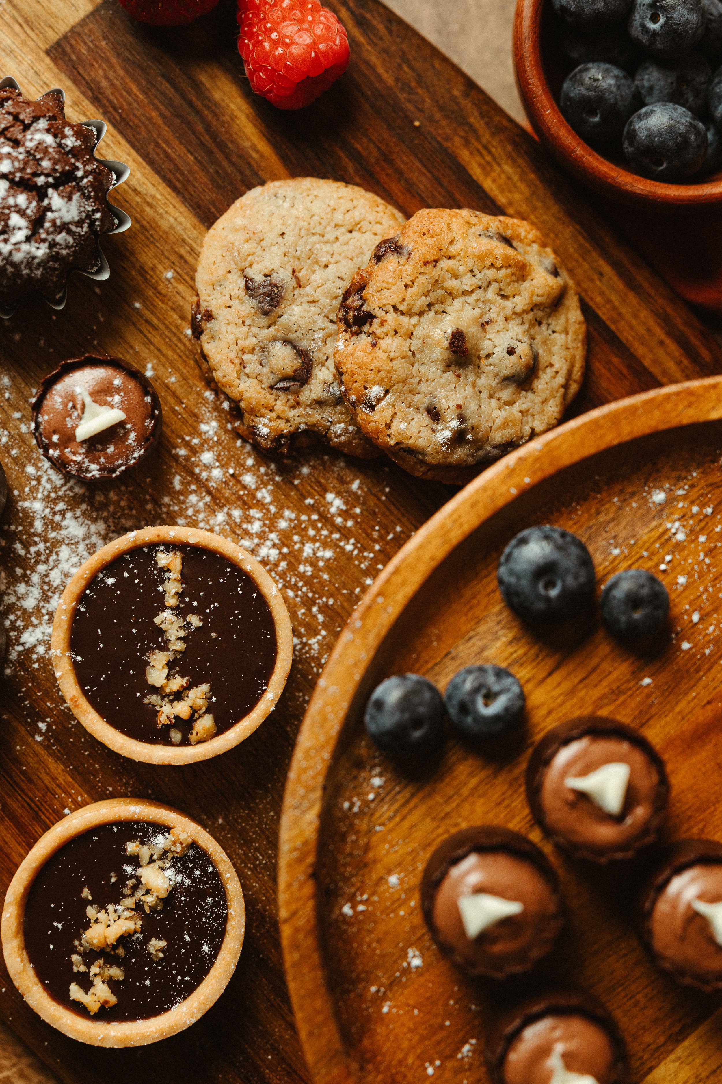 Assorted cookies, blueberries in a wooden bowl, raspberries, and chocolate tarts on a wooden tray.