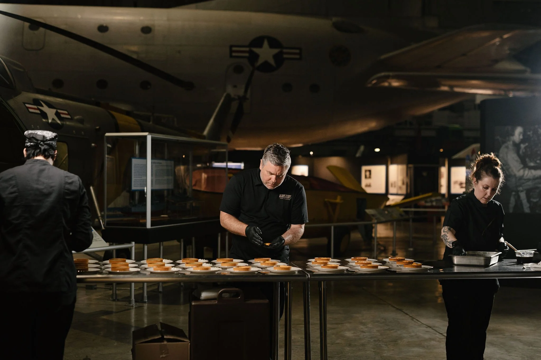 Museum exhibit with a large aircraft overhead, and three people in black uniforms preparing food or snacks at a table.