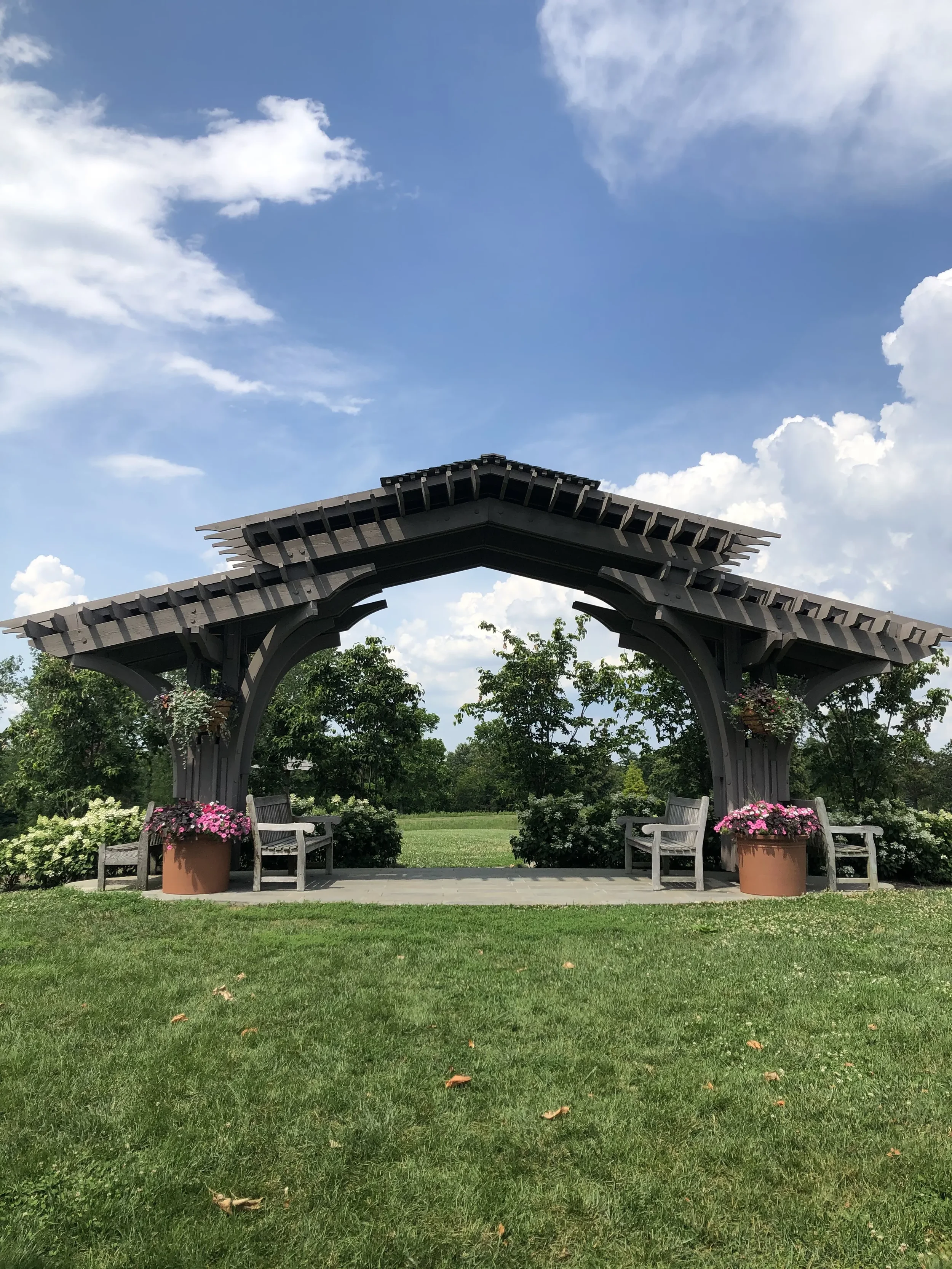 A decorative wooden pavilion with benches and flower pots in a park on a sunny day with a blue sky and some clouds.