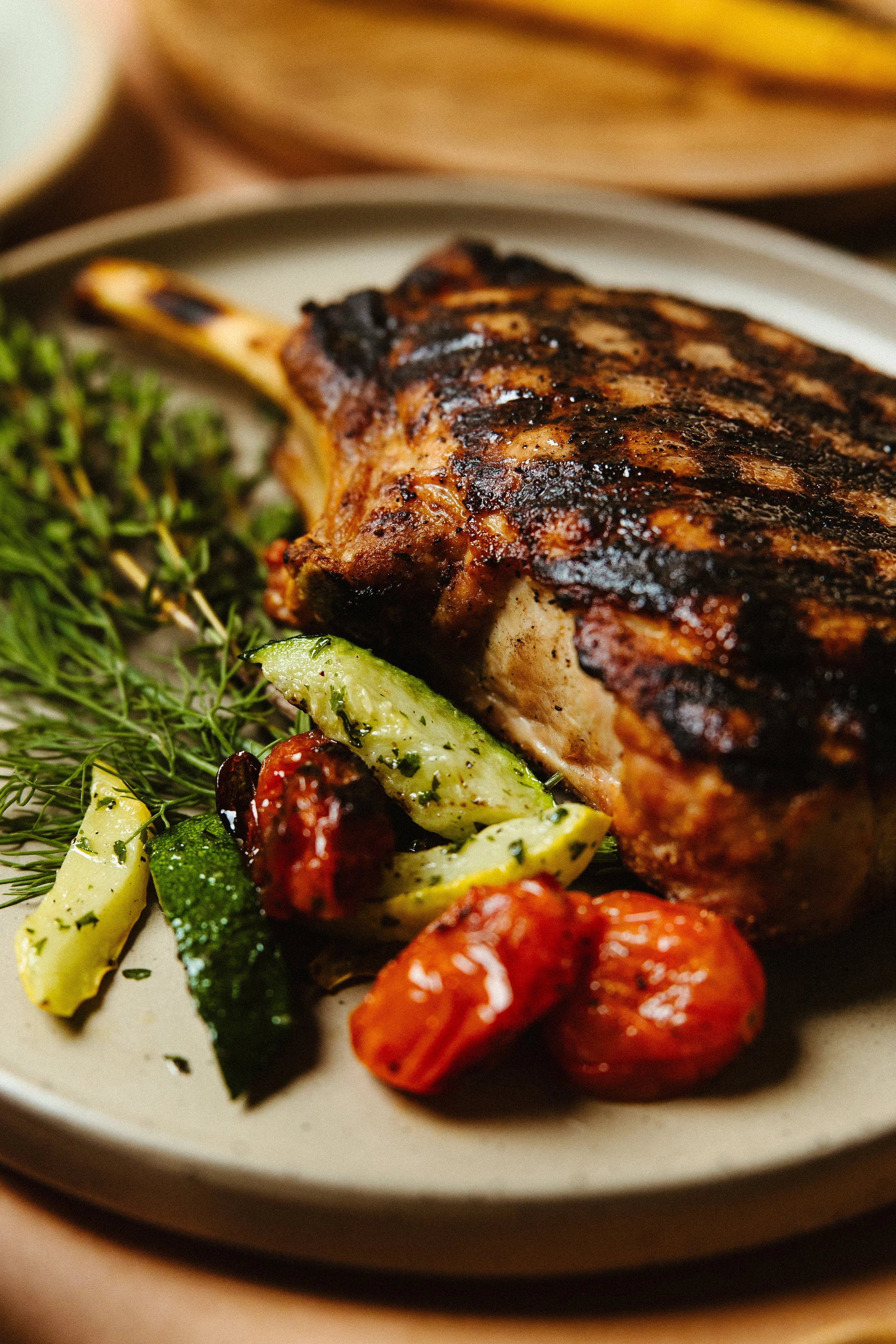 Close-up of a grilled pork chop with char marks, garnished with fresh herbs, and served with roasted cherry tomatoes and zucchini.