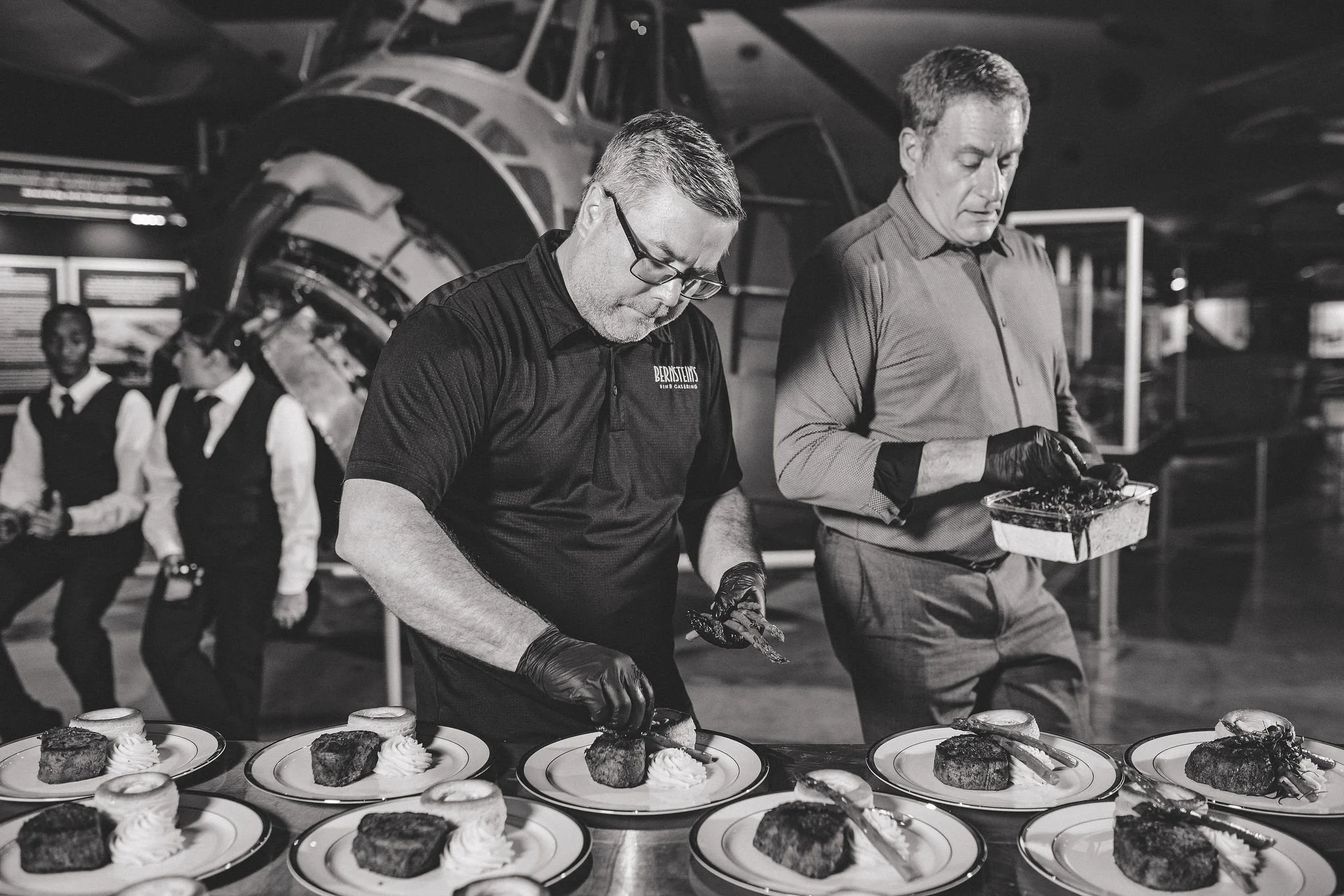 Two men in aprons and black gloves prepare plates of food on a counter, with a background of an aircraft and three waitstaff standing in line.
