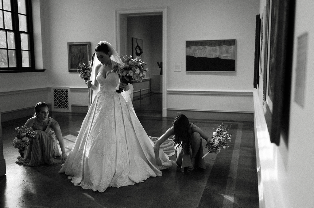 A bride in a wedding gown holding a bouquet, sitting on her wedding day with two bridesmaids adjusting her dress in an art gallery or museum room.