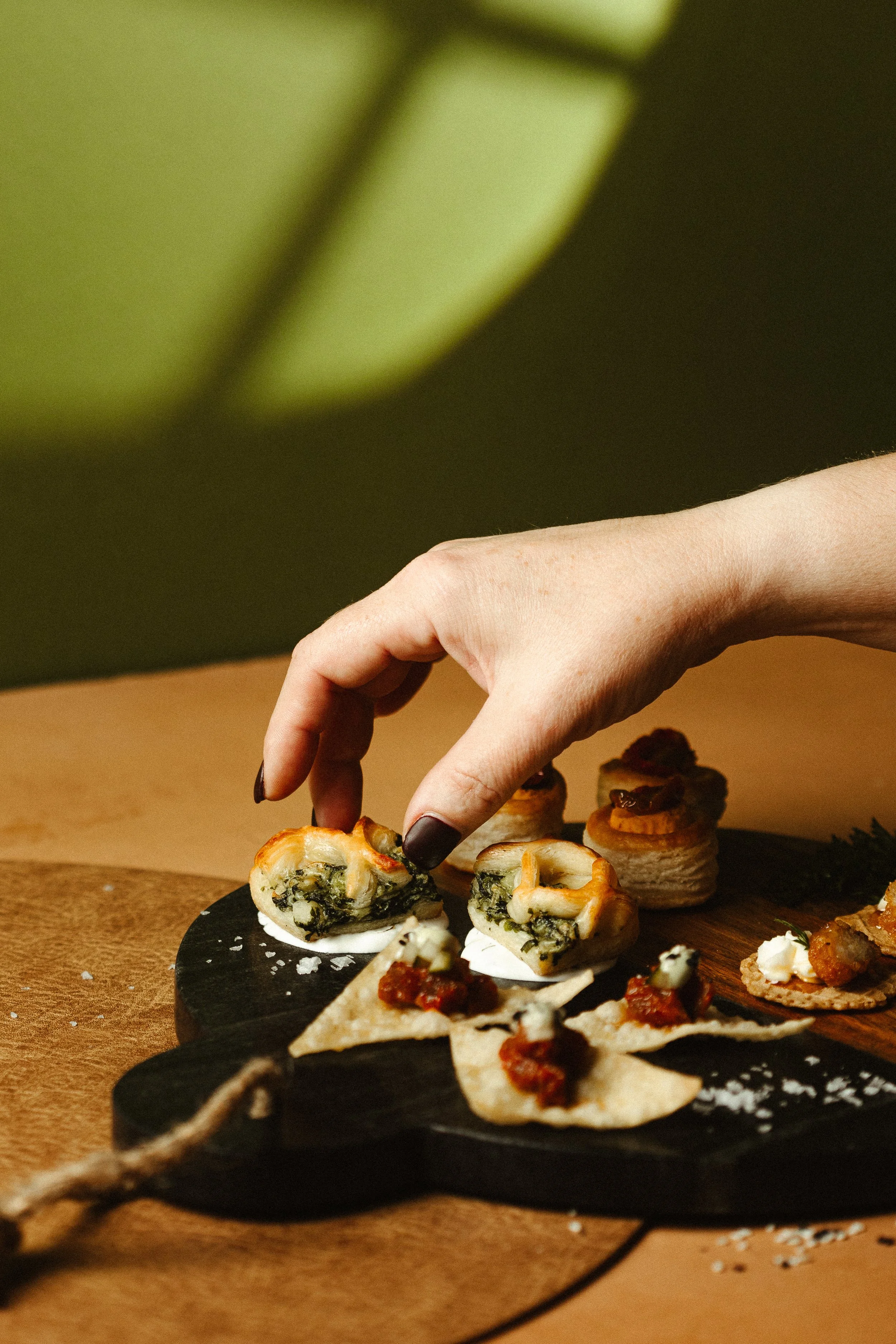 Close-up of a hand reaching for mini savory pastries on black slate serving platter, with additional appetizers in the background.