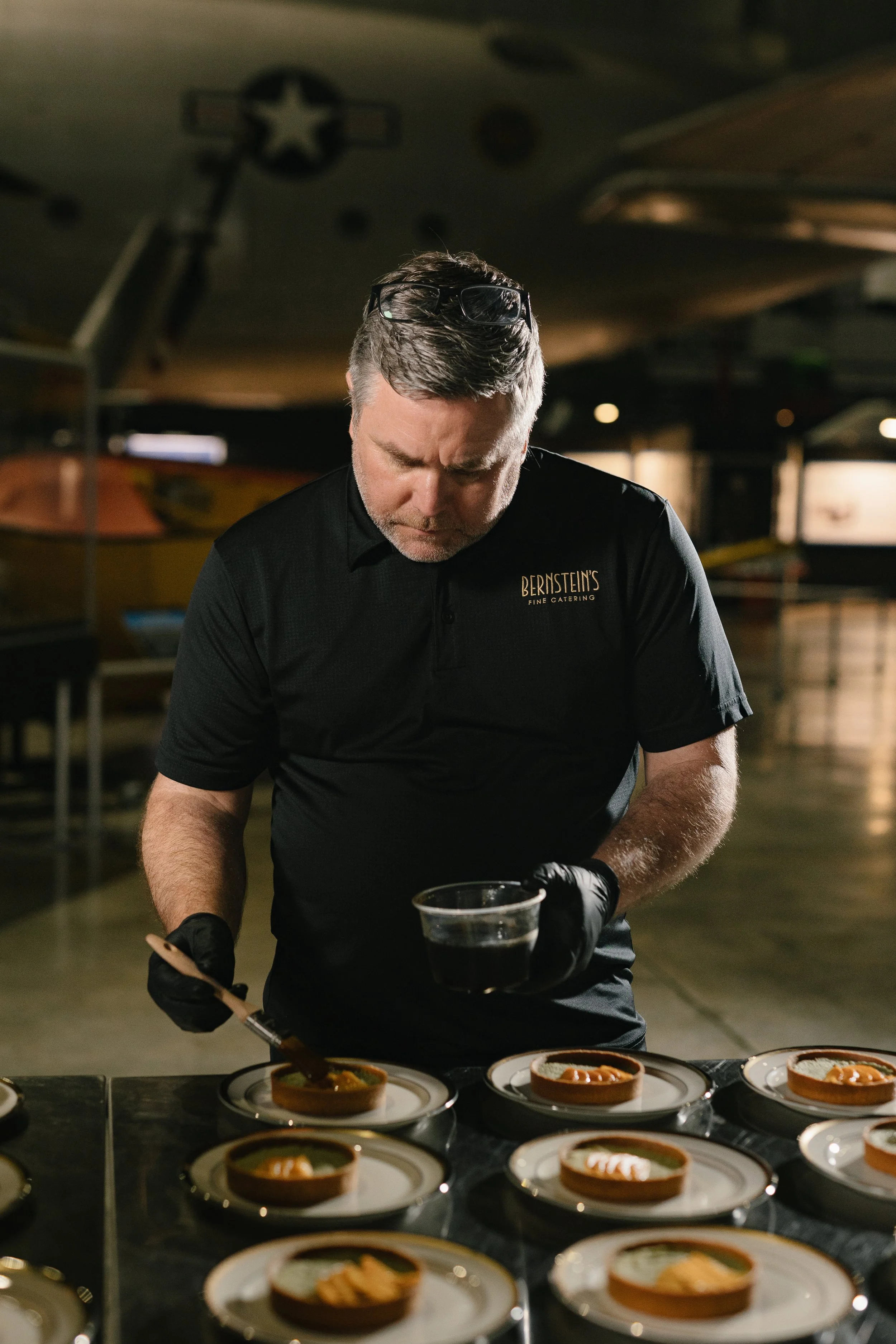 A man wearing a black shirt with 'Bernstein's Fine Catering' embroidered, is preparing food in a dimly lit catering area. He is handling a container, with several plates of food arranged on a table in front of him.