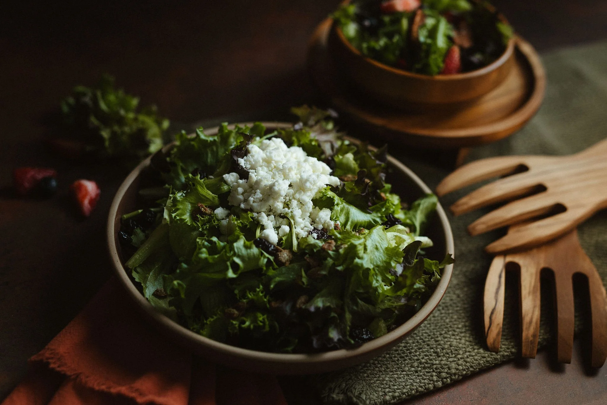 Bowl of mixed greens salad topped with crumbled cheese, with a wooden salad spinner and a small wooden bowl of berries in the background.