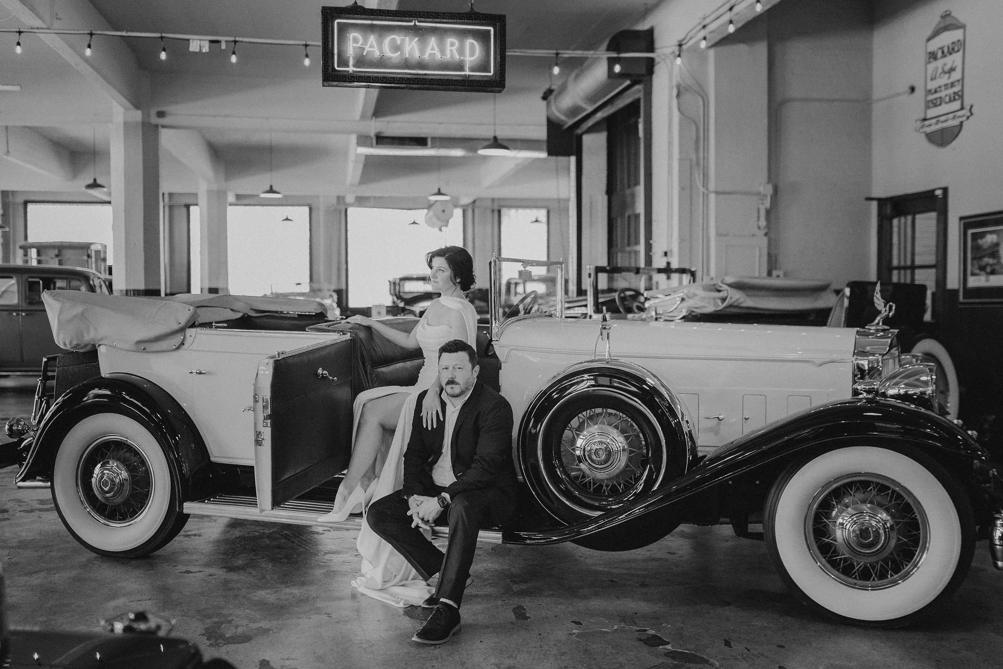 A black-and-white photo of a vintage car inside a showroom, with a woman and a man posing by the car, and a neon 'Packard' sign hanging above.