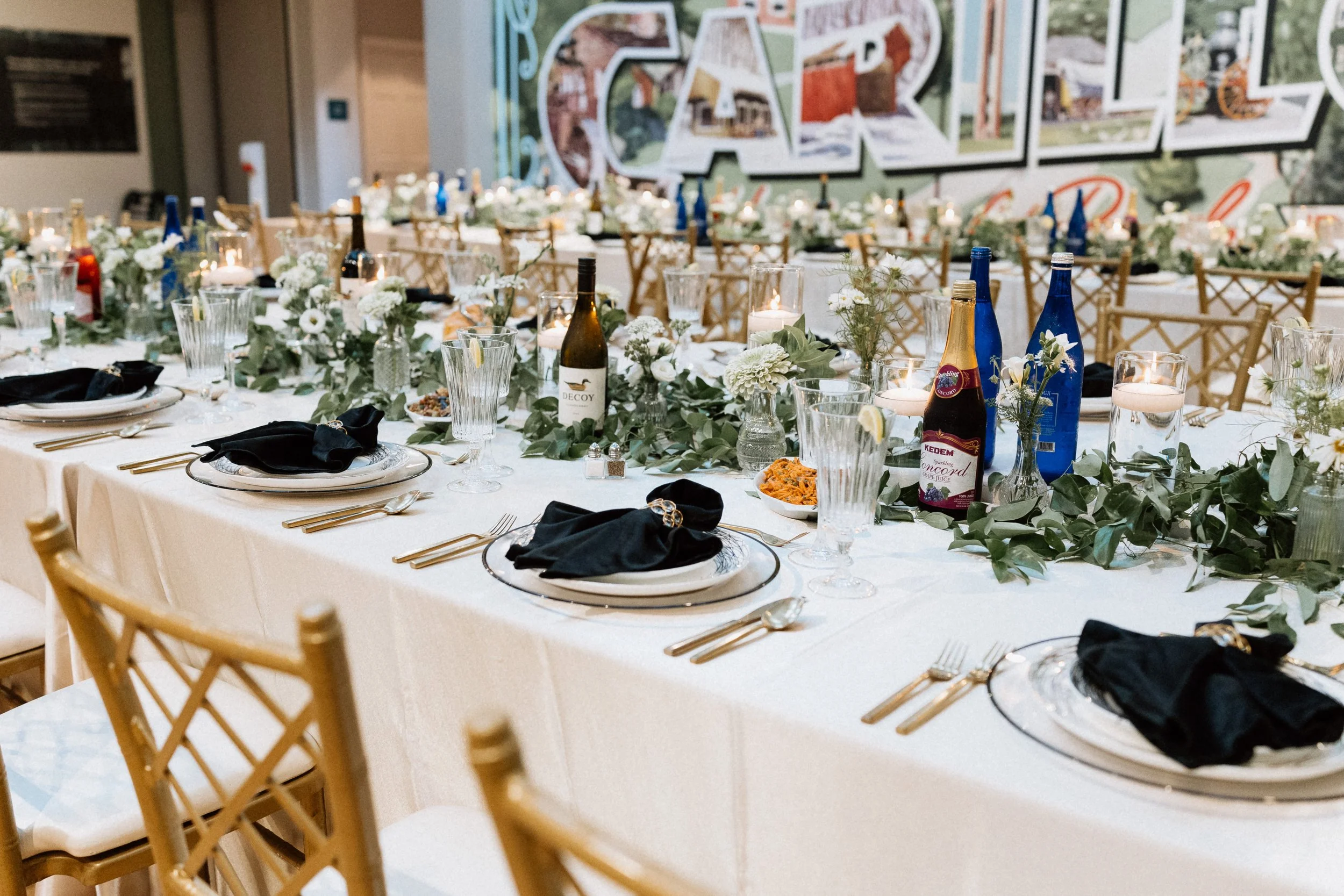 A banquet table decorated with white flowers, greenery, candles, and bottles of beverages, set for a formal event or celebration with gold-colored chairs and a large colorful wall mural in the background.