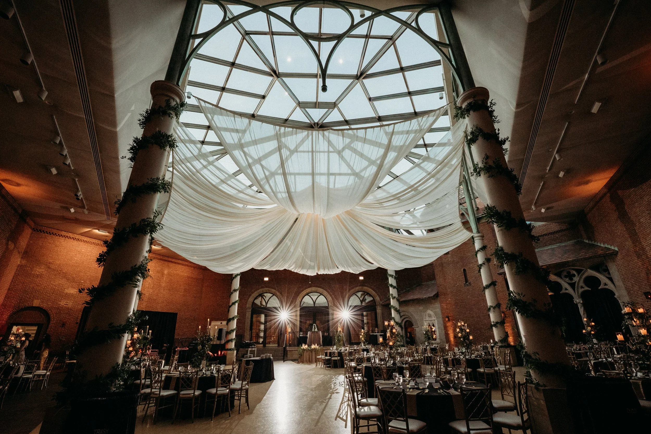 Elegant event space with high glass ceiling, draped white fabric, tall columns with greenery, circular tables set for a formal event, warm lighting, brick walls, and a dance floor in the center.