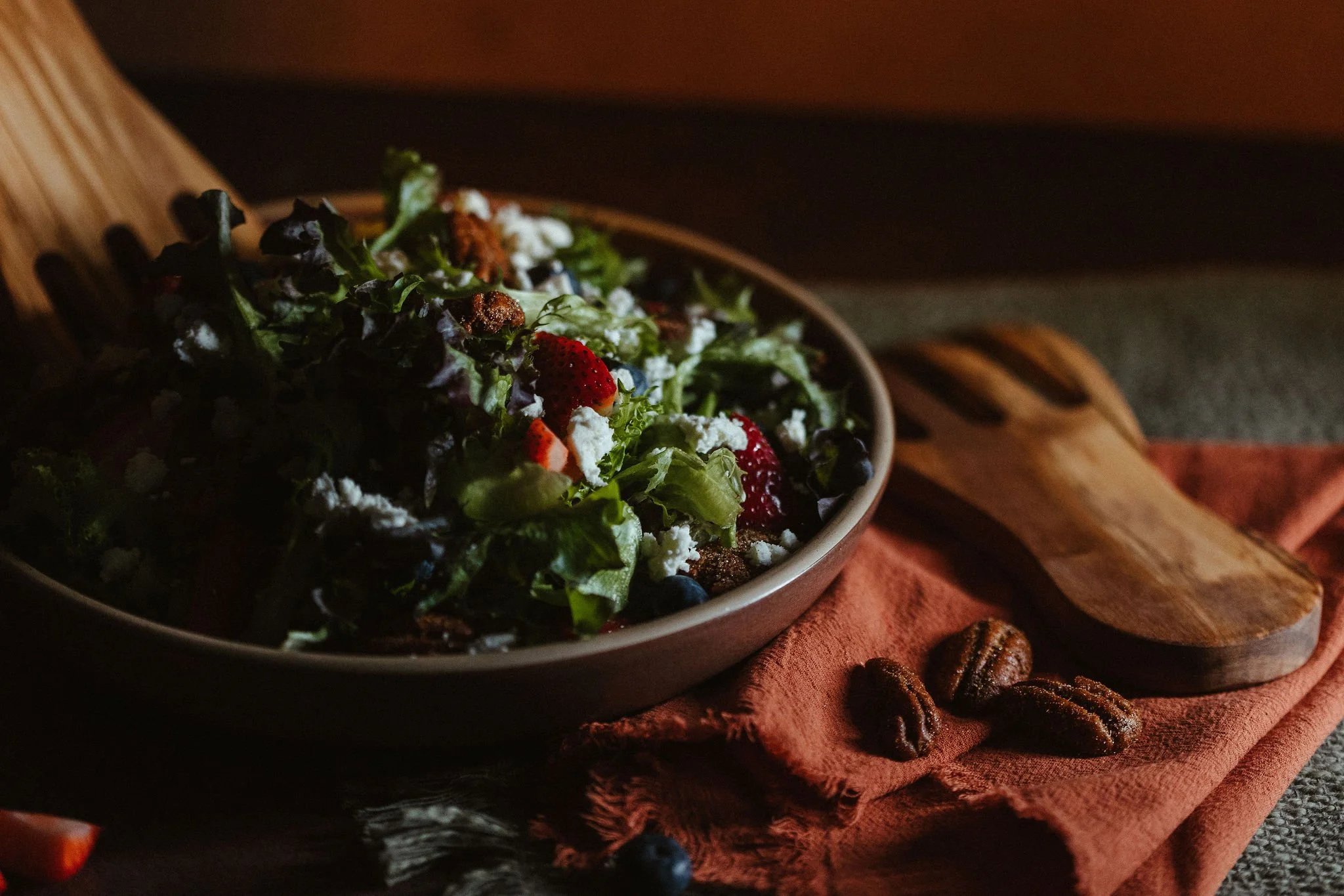 A bowl of mixed salad with strawberries, blueberries, feta cheese, and greens on a wooden serving board, with a rust-colored cloth napkin and cinnamon sticks in the background, on a gray surface.