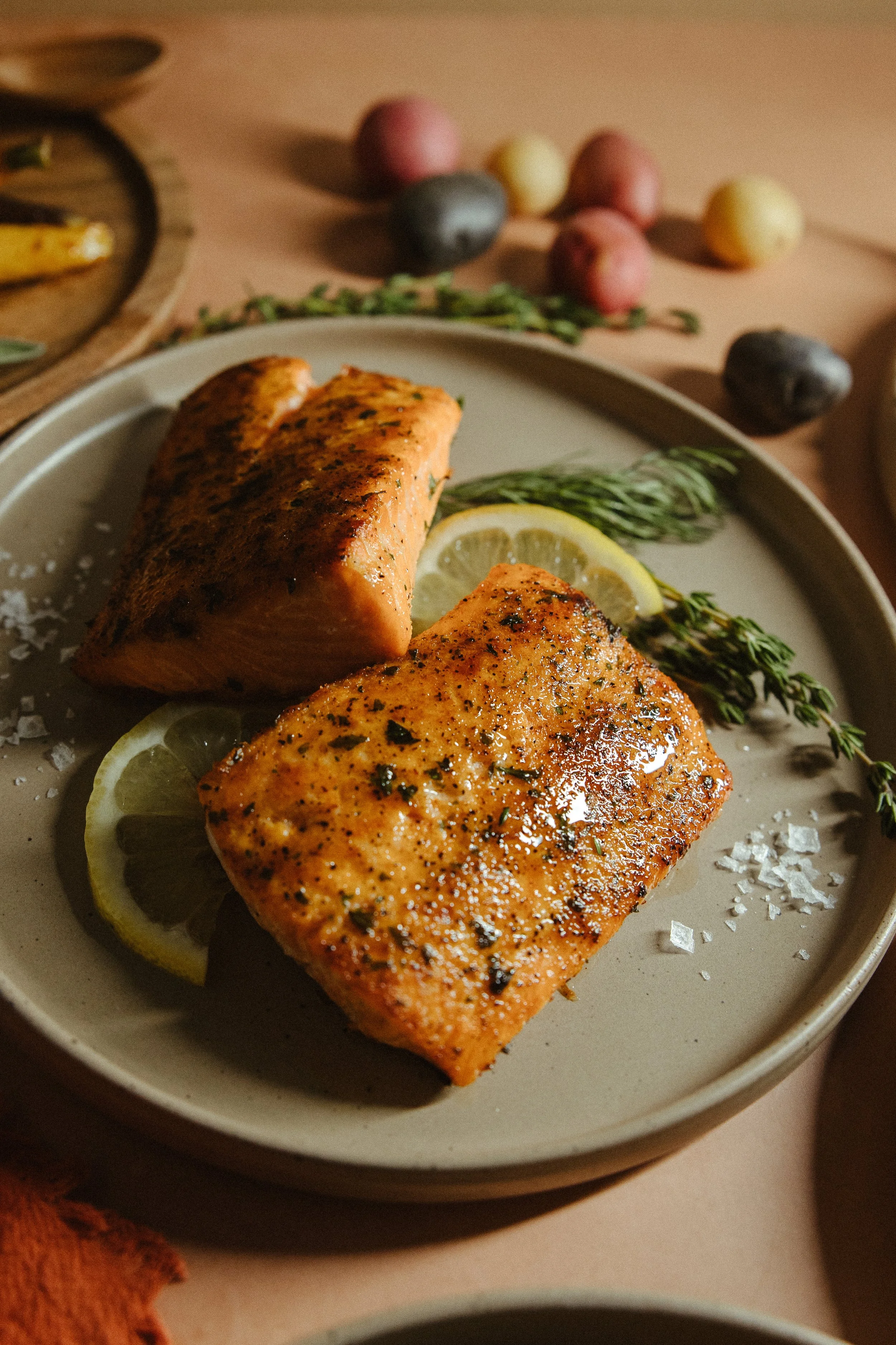 A plate with seasoned baked salmon, garnished with lemon slices and fresh herbs, on a beige table with colorful small potatoes and a sprig of thyme in the background.