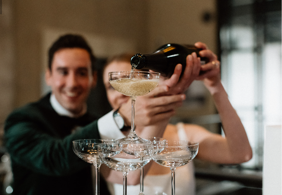 A man in a tuxedo pouring champagne into a champagne coupe held by a woman, with a pyramid of other coupe glasses in front of them.