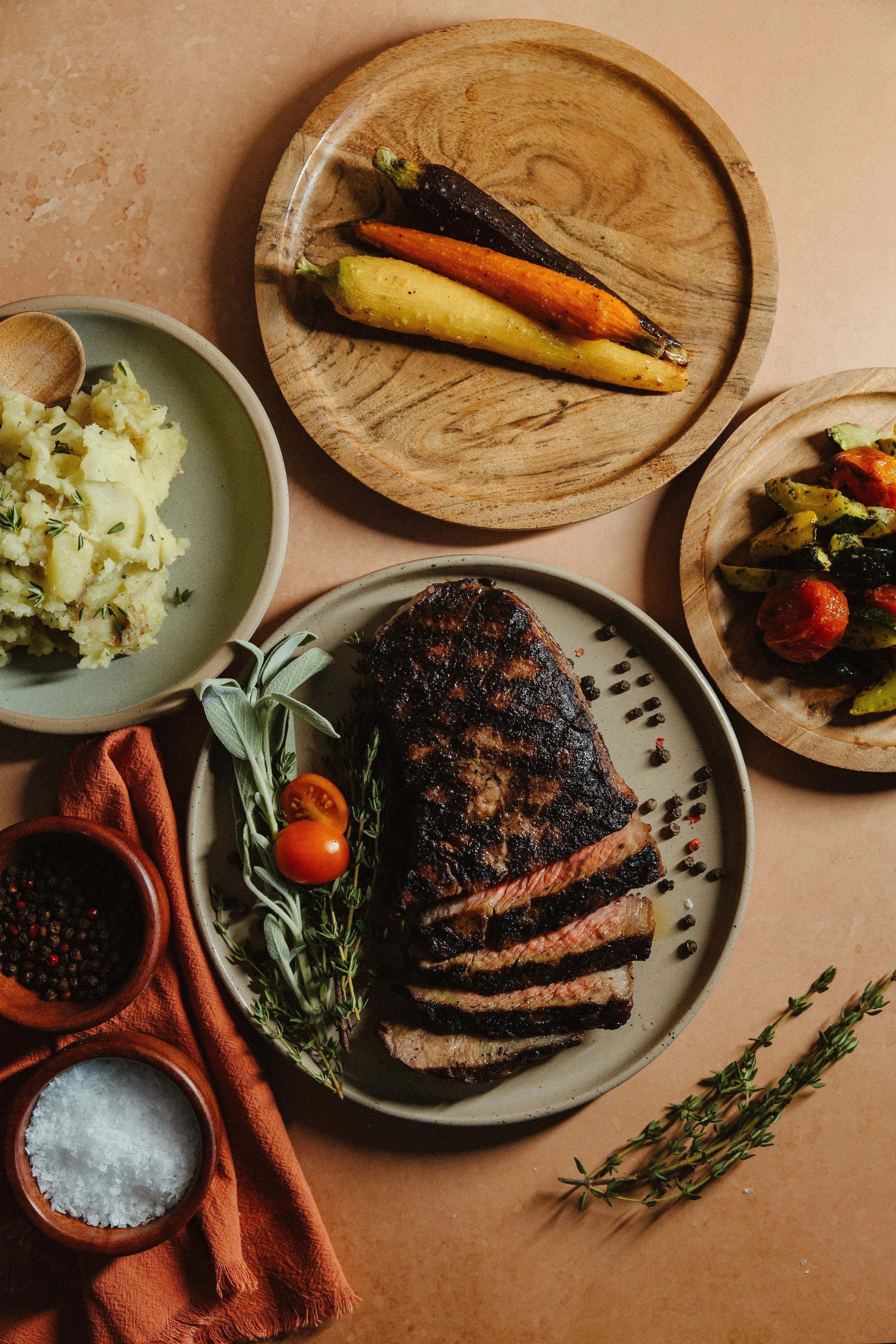 Sliced medium-rare steak garnished with herbs, surrounded by tomatoes, rosemary, and salt, with side dishes of mashed potatoes and roasted vegetables on wooden plates.