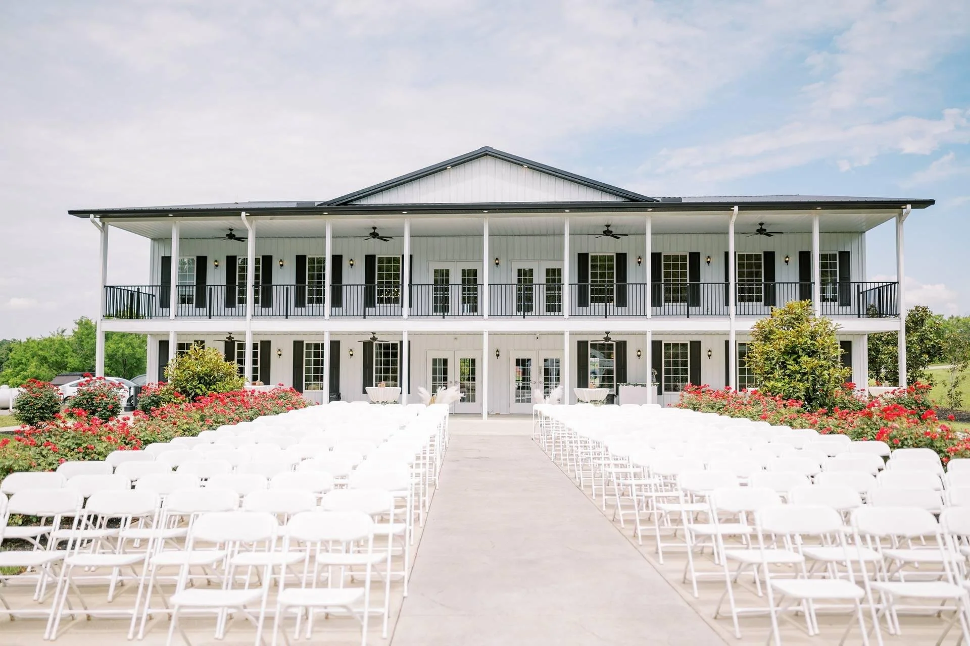 Outdoor wedding setup with white chairs arranged in rows, floral decorations, and a large white building with balconies and ceiling fans in the background.