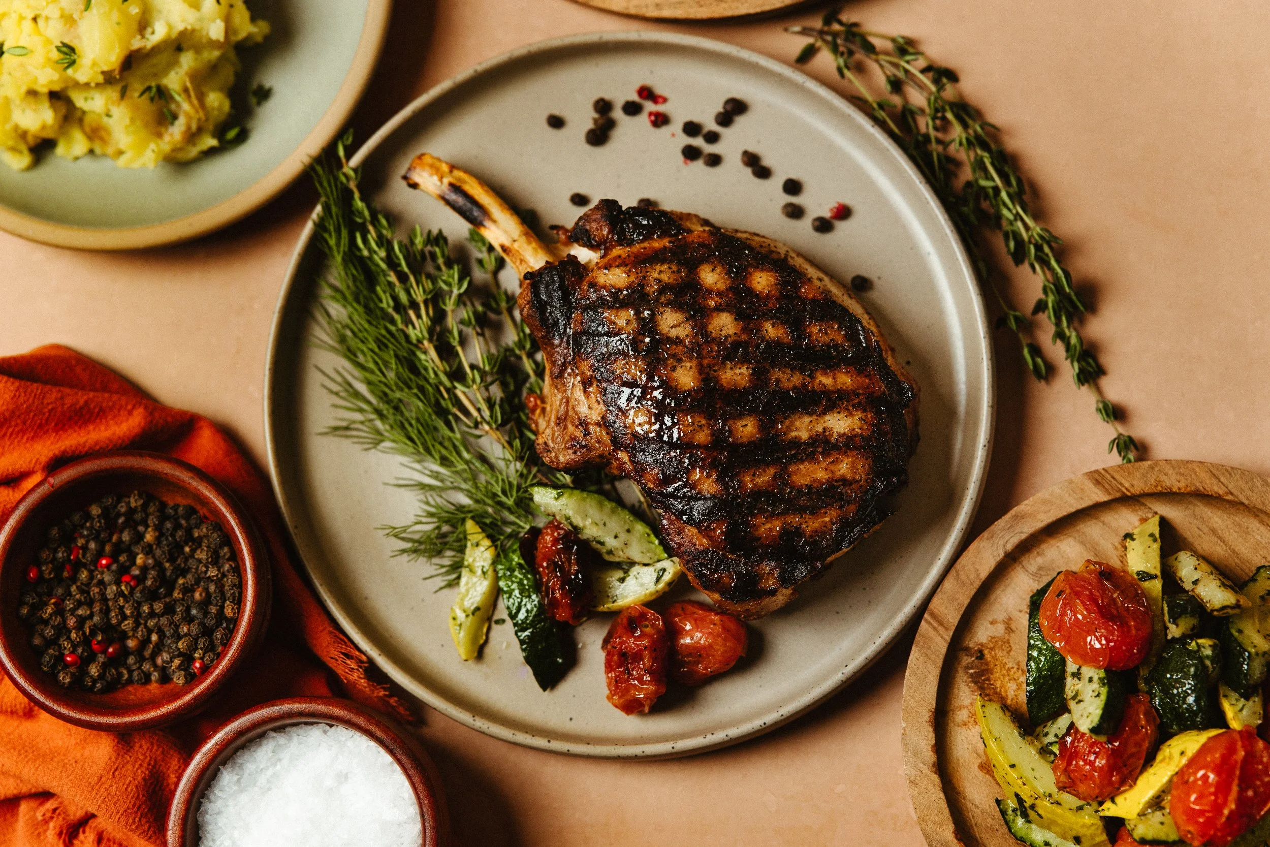 A plated grilled pork chop garnished with herbs, with roasted vegetables and a side of salad. Surrounding bowls contain peppercorns, salt, and black pepper.