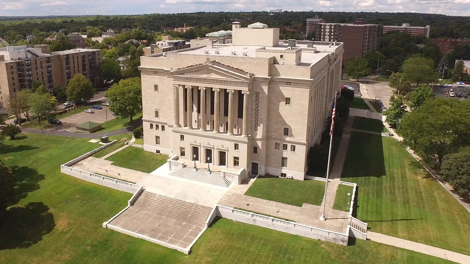 An aerial view of a historic white stone building with classical columns labeled 'Masonic Temple,' surrounded by green lawns, trees, and parking lots in an urban area.