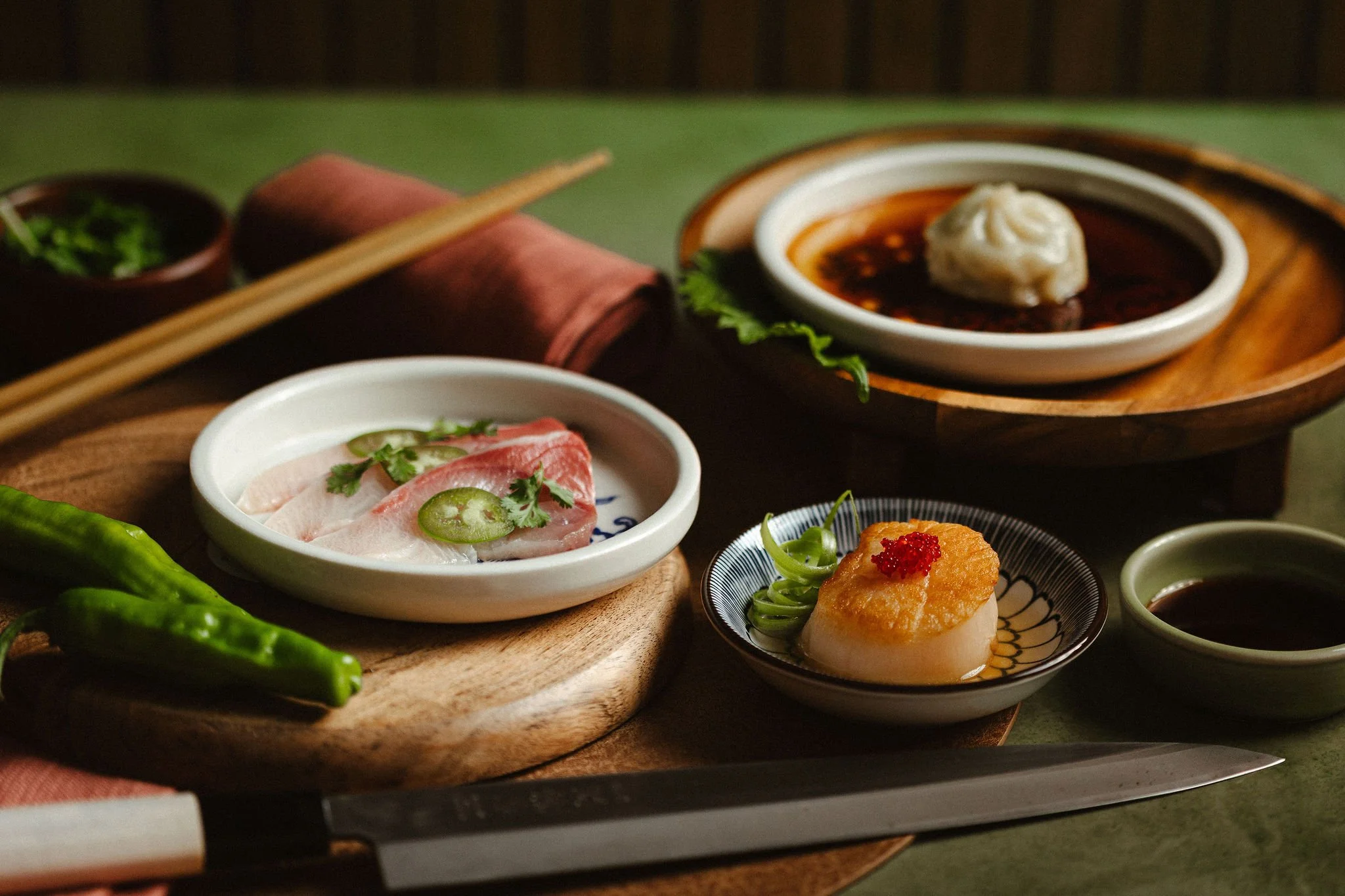 Assorted Asian dishes including sliced fish sashimi with green chili slices and cilantro, a bowl of dark dipping sauce with a dumpling on top, and a small serving of fried food with garnish, all arranged on a wooden serving board.