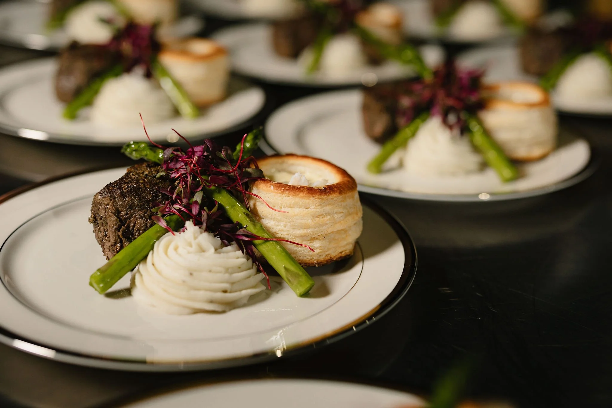 Elegant plated appetizer featuring a cooked beef tenderloin, a pastry shell filled with creamy filling, a swirl of mashed potatoes, green beans, and microgreens, all presented on a white and gold-rimmed plate.