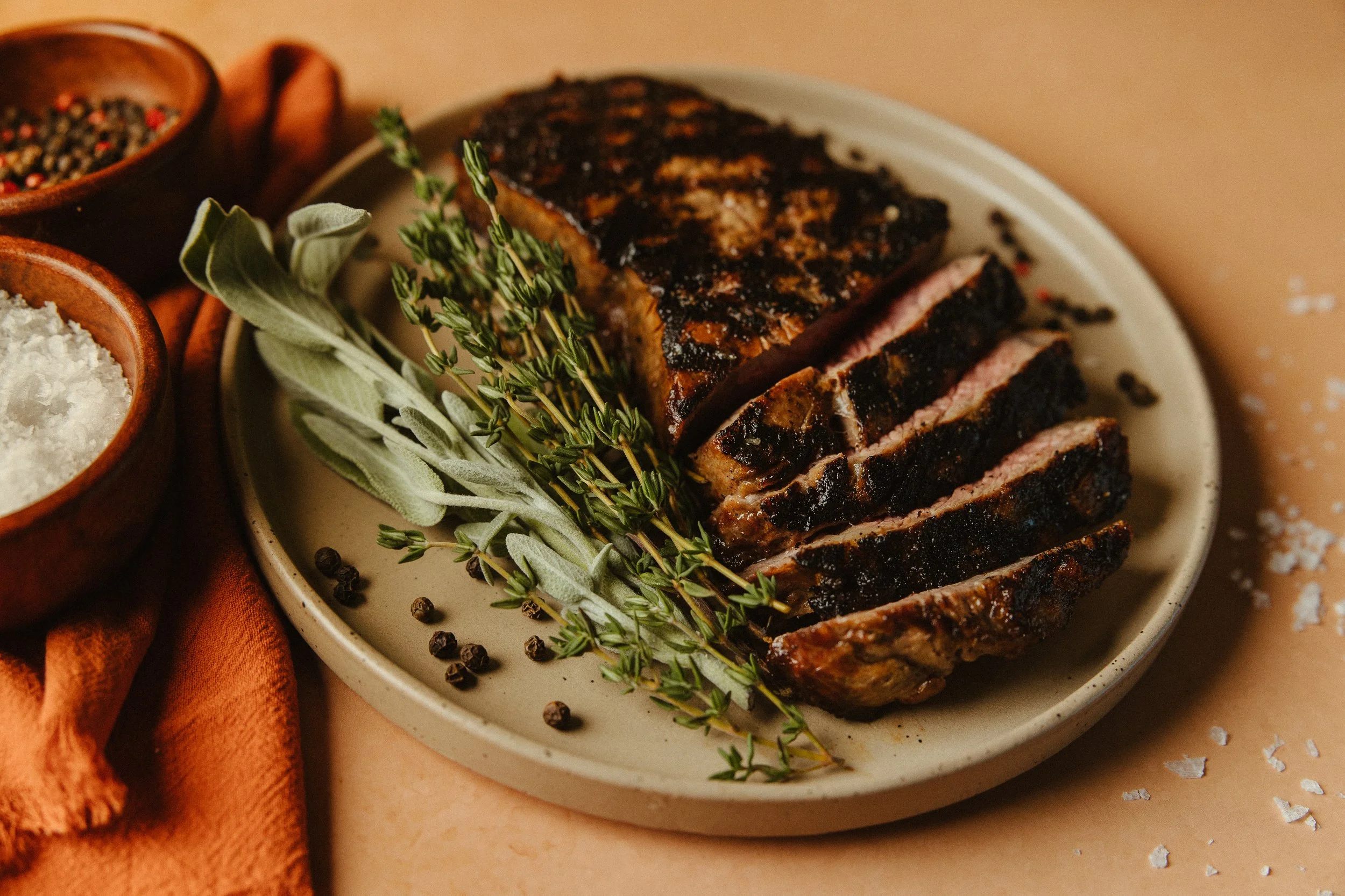 Slices of grilled steak on a white plate garnished with fresh herbs, with small bowls of salt, pepper, and seasoning nearby.