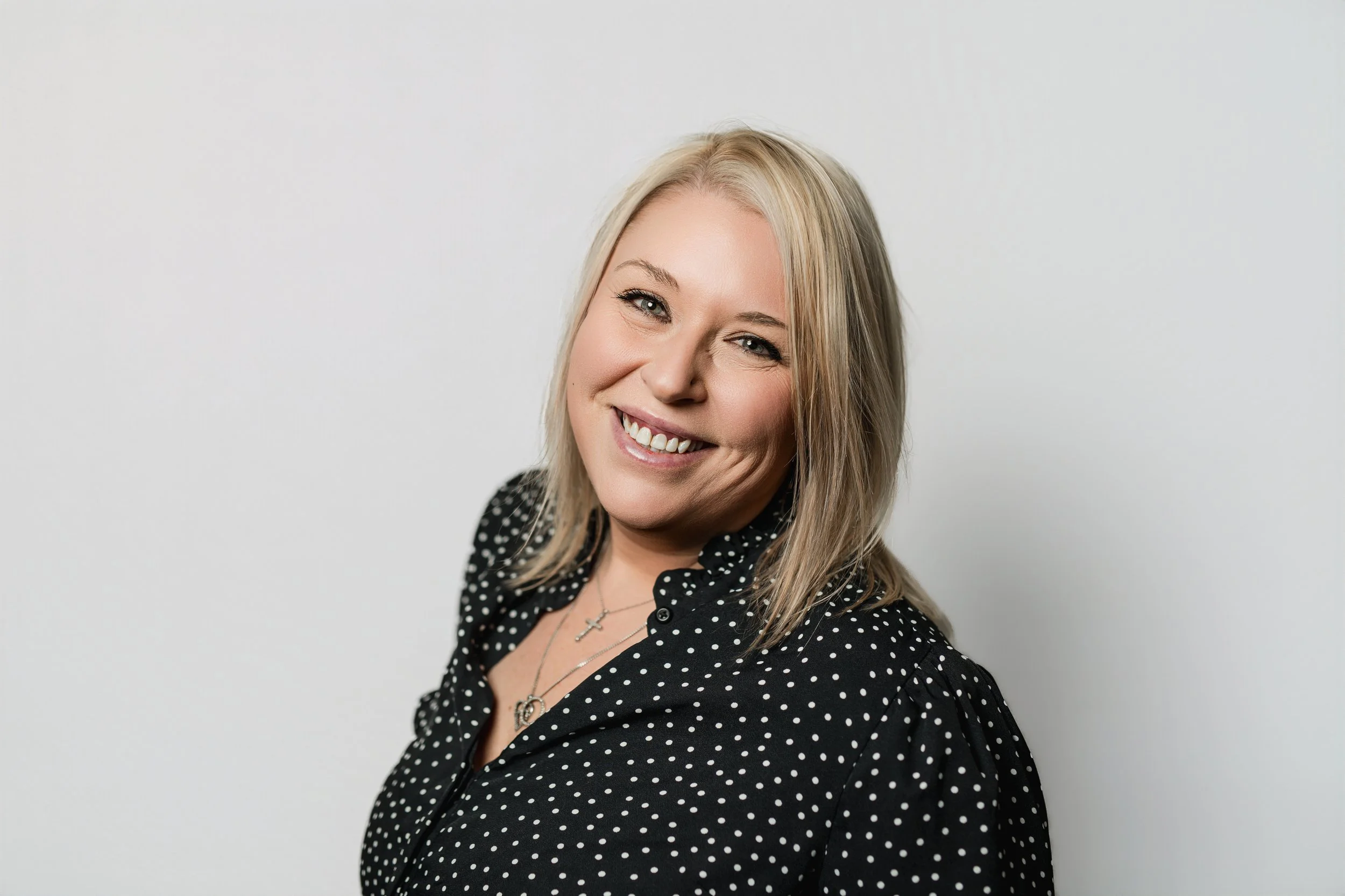 Portrait of a smiling blonde woman with blue eyes, wearing a black polka dot blouse and jewelry, standing against a plain white background.
