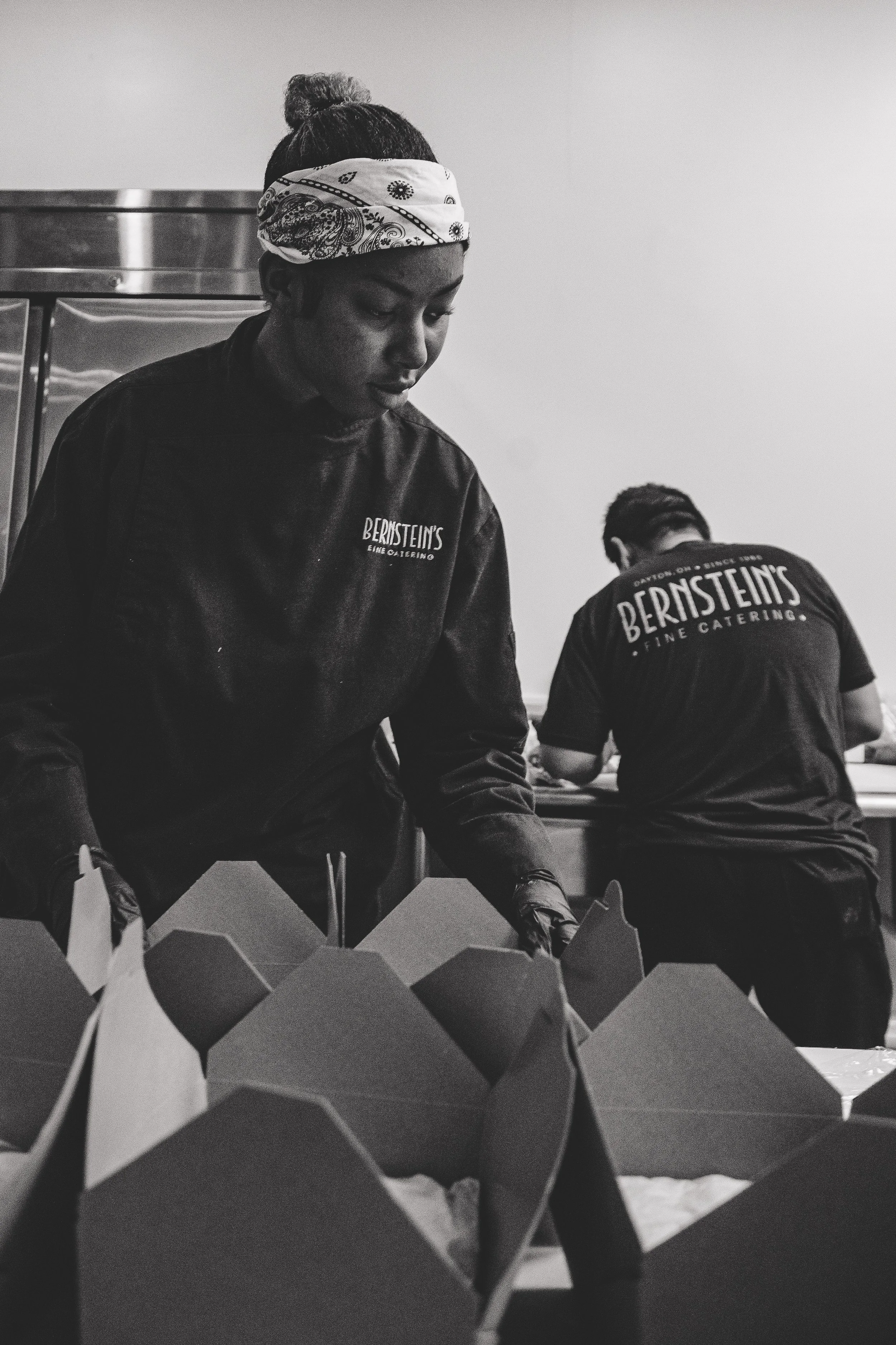 Black and white photo of two people working at a counter, one wearing a bandana and a chef's jacket with 'Bernstein's' logo, the other with 'Bernstein's' logo on their shirt.