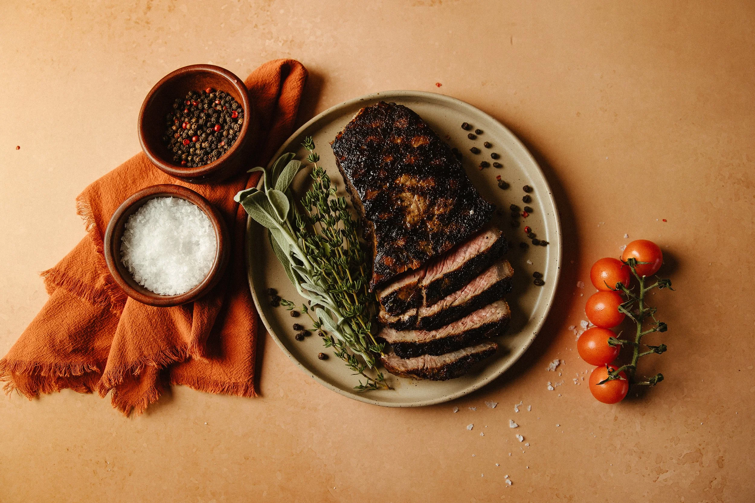 Slices of grilled steak on a plate garnished with sage and thyme, with bowls of salt and pepper nearby, on an orange cloth, with cherry tomatoes on the vine to the side.