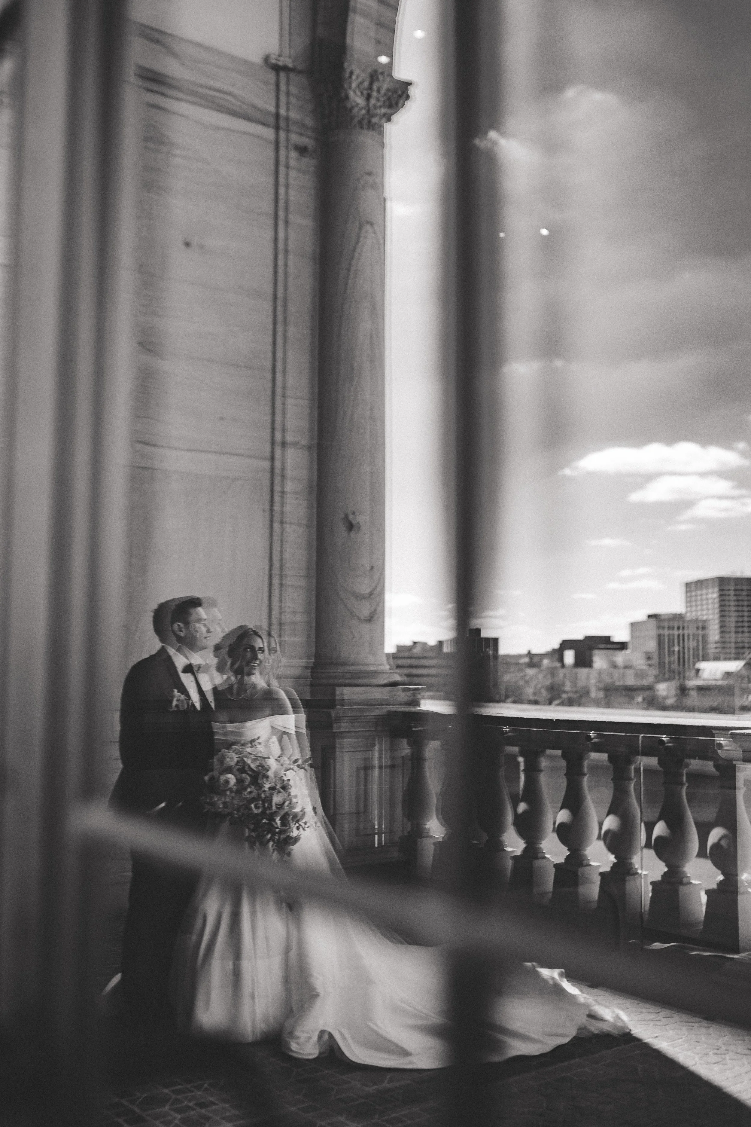 Black and white photo of a bride and groom standing on a balcony, with a cityscape in the background. The bride is holding a bouquet of flowers, and they appear to be smiling.