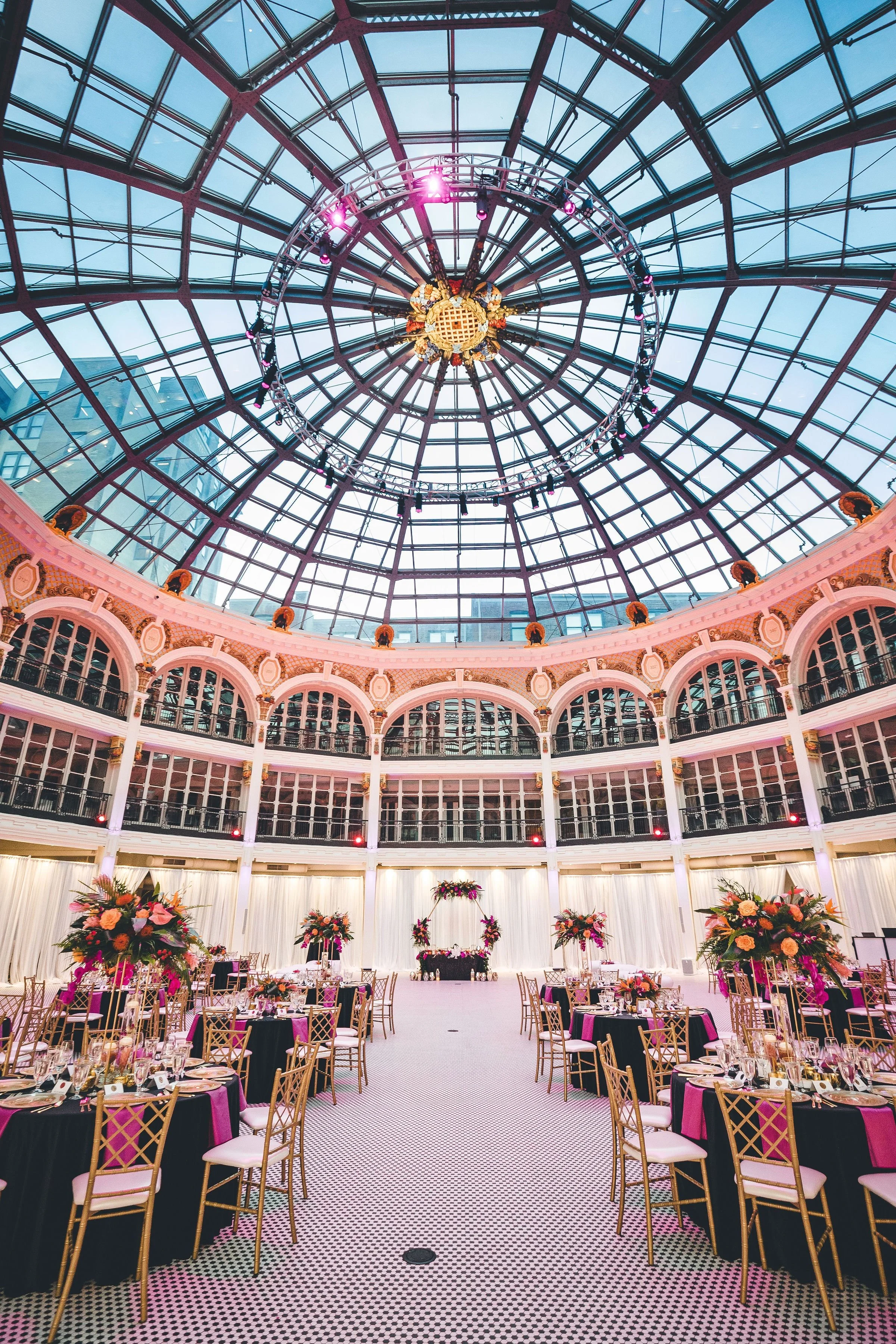 Elegant event space with a glass dome ceiling, pink and gold floral arrangements, and round tables with black tablecloths and gold chairs, set for a formal occasion.