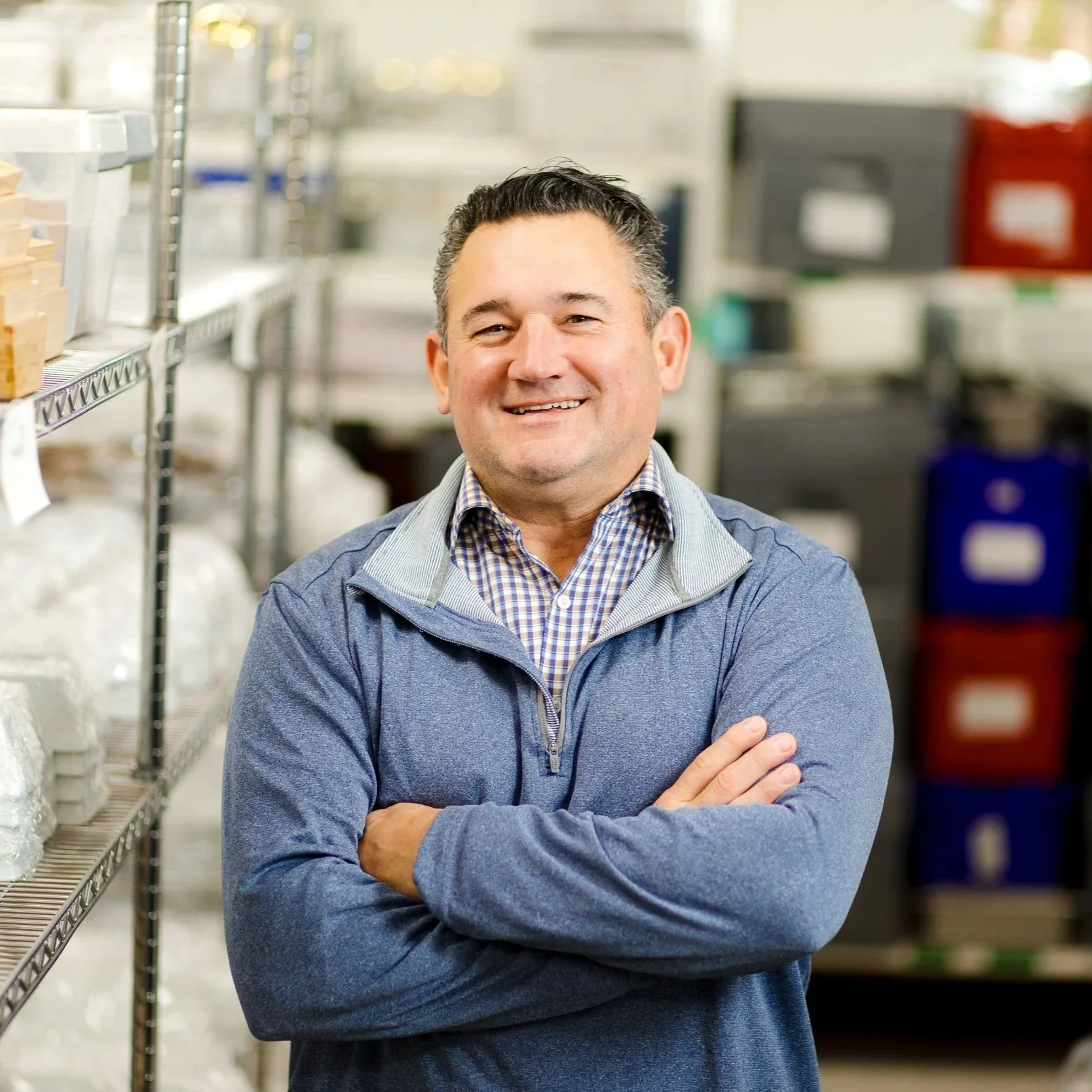 Man smiling with arms crossed in a storage room or warehouse with shelves of supplies behind him.