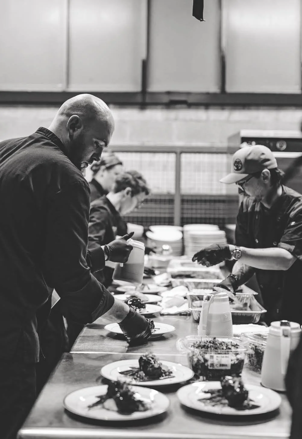 Black and white photo of chefs preparing food at a busy kitchen counter with plates and various ingredients.