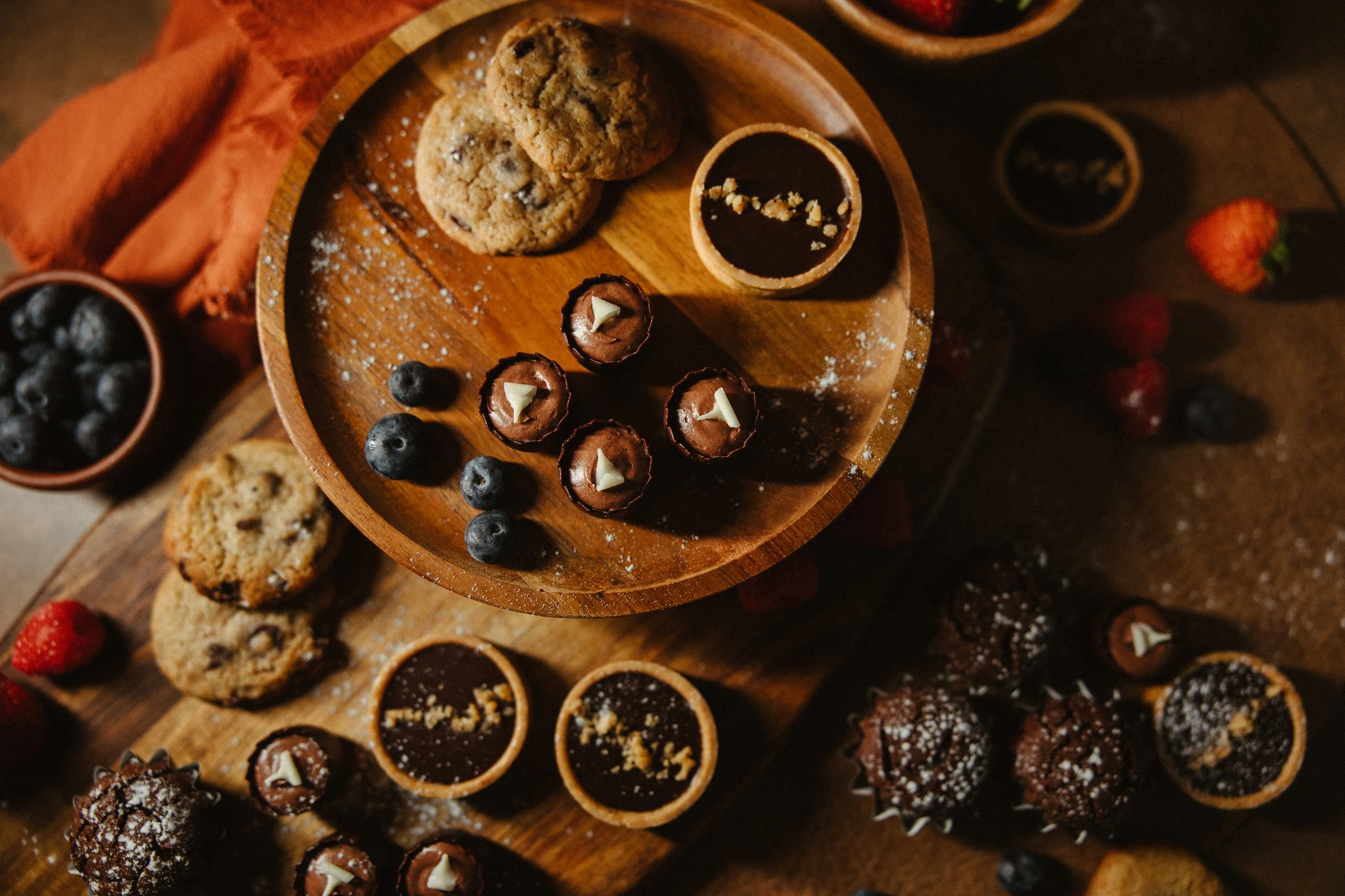 Assorted cookies and chocolate cups on wooden serving trays with strawberries and blueberries nearby.