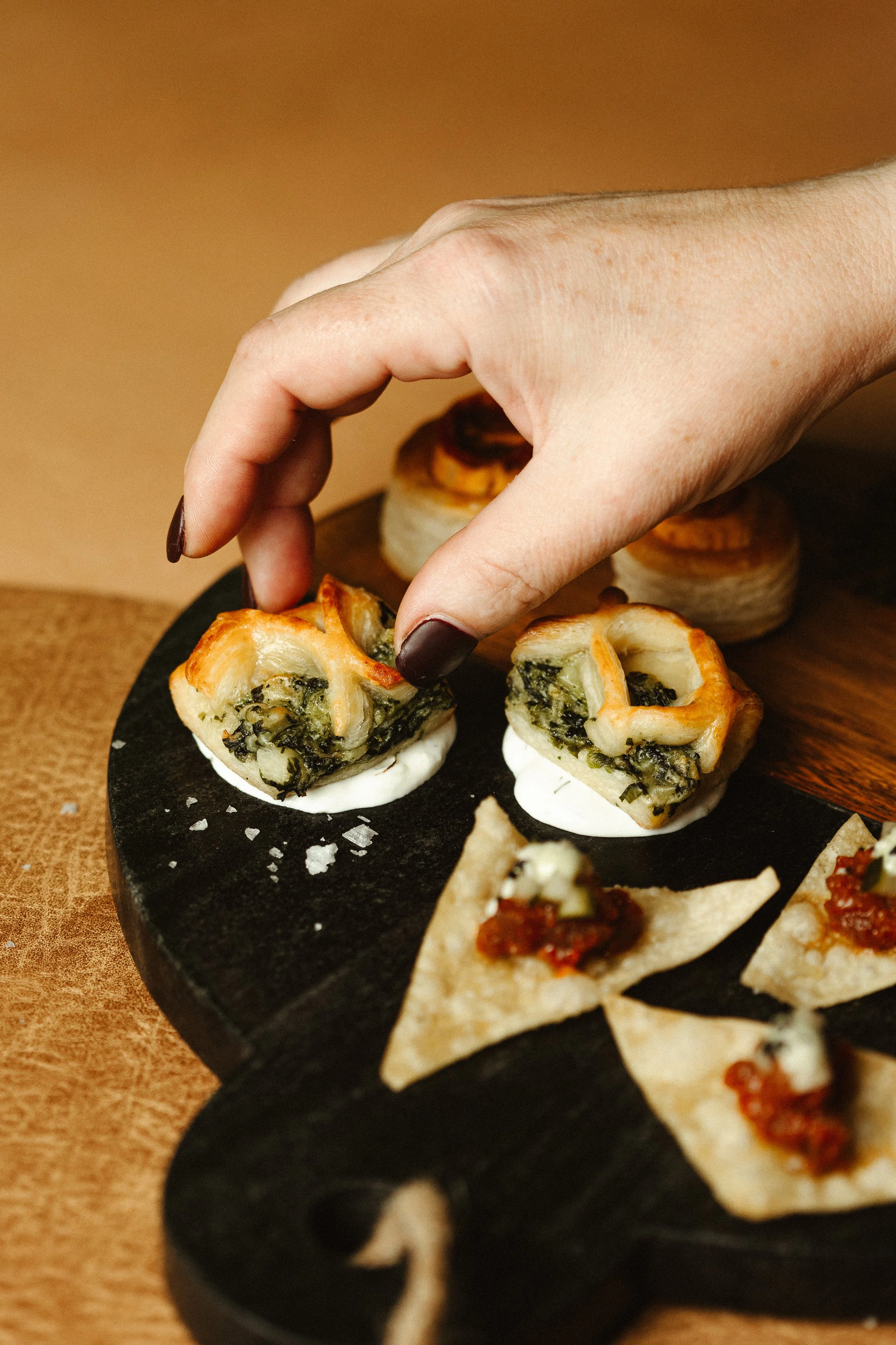 Close-up of stuffed pastry appetizers on a black serving platter, with a hand picking up one piece. The platter also has small pieces of bread or crackers with toppings.