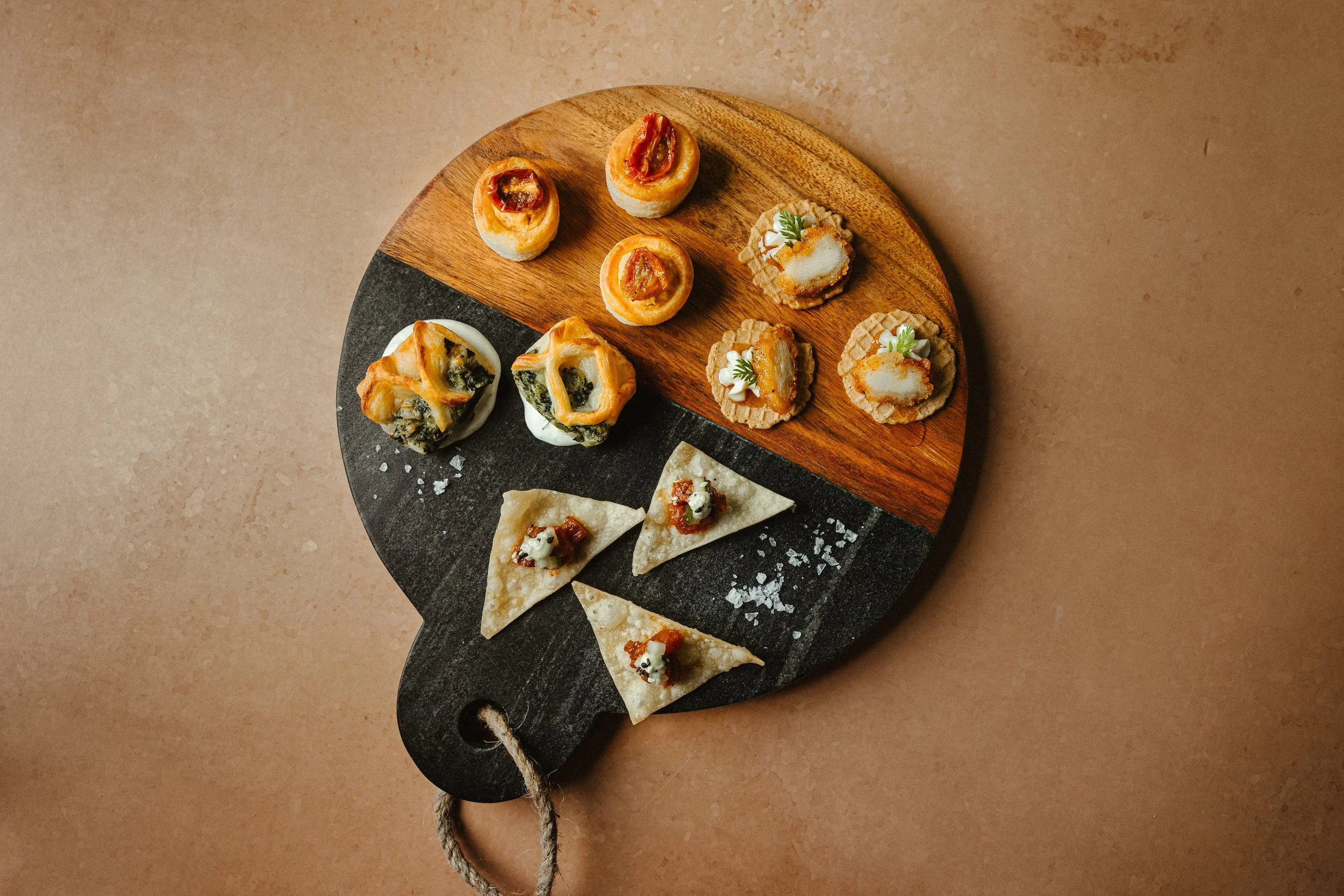 Assorted mini appetizers on a round wooden and slate serving board, including small slices of layered pastry with toppings, mini tostadas topped with cheese and sauce, and bite-sized open-faced sandwiches with greens.