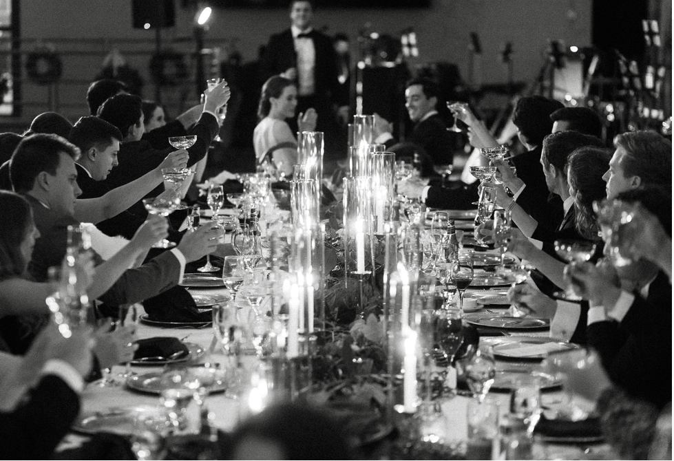 People in formal attire at a banquet table, raising glasses in a toast, with candles and elegant table settings, in a black-and-white photo.
