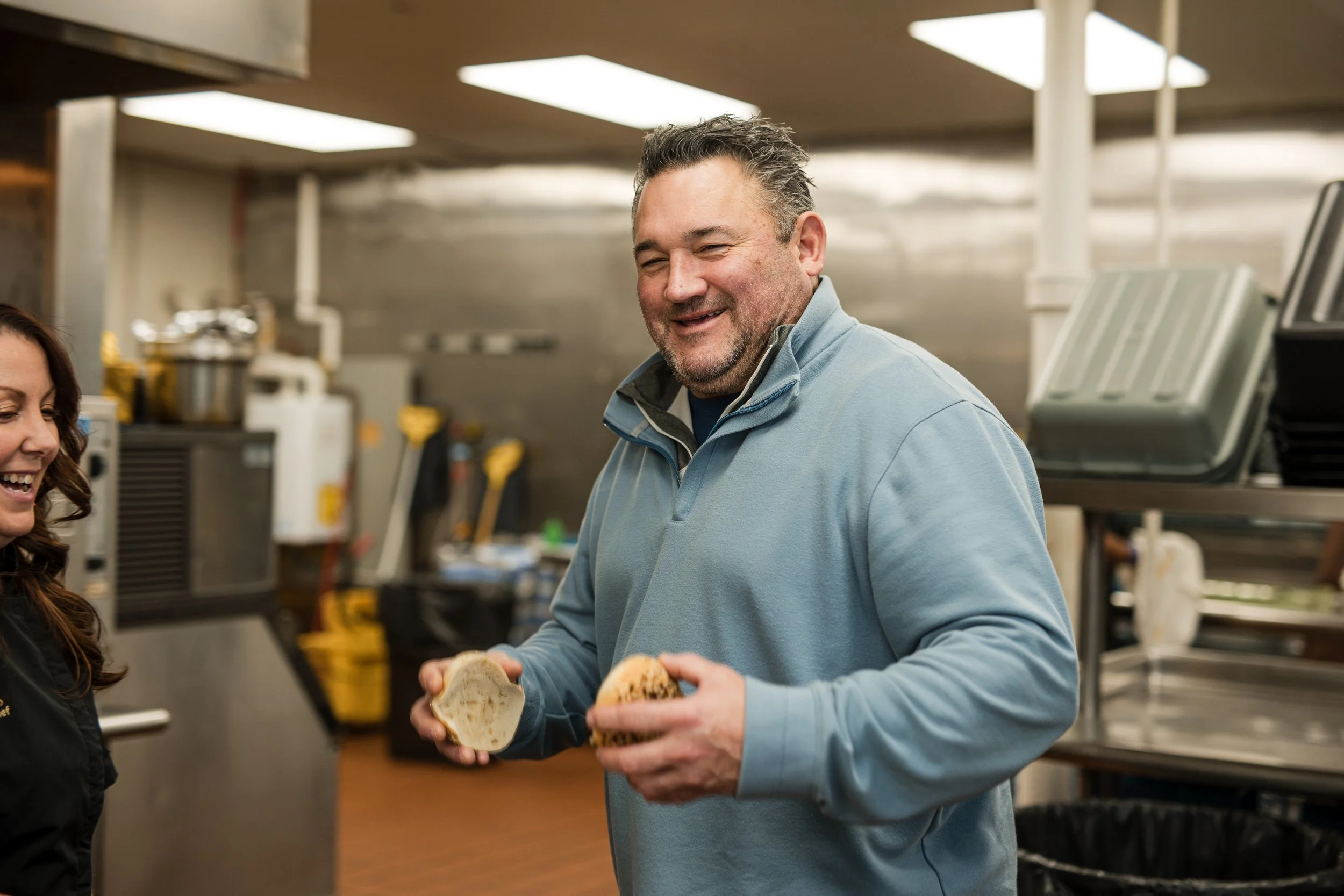A man in a light blue jacket holding bread buns and talking, in a commercial kitchen.
