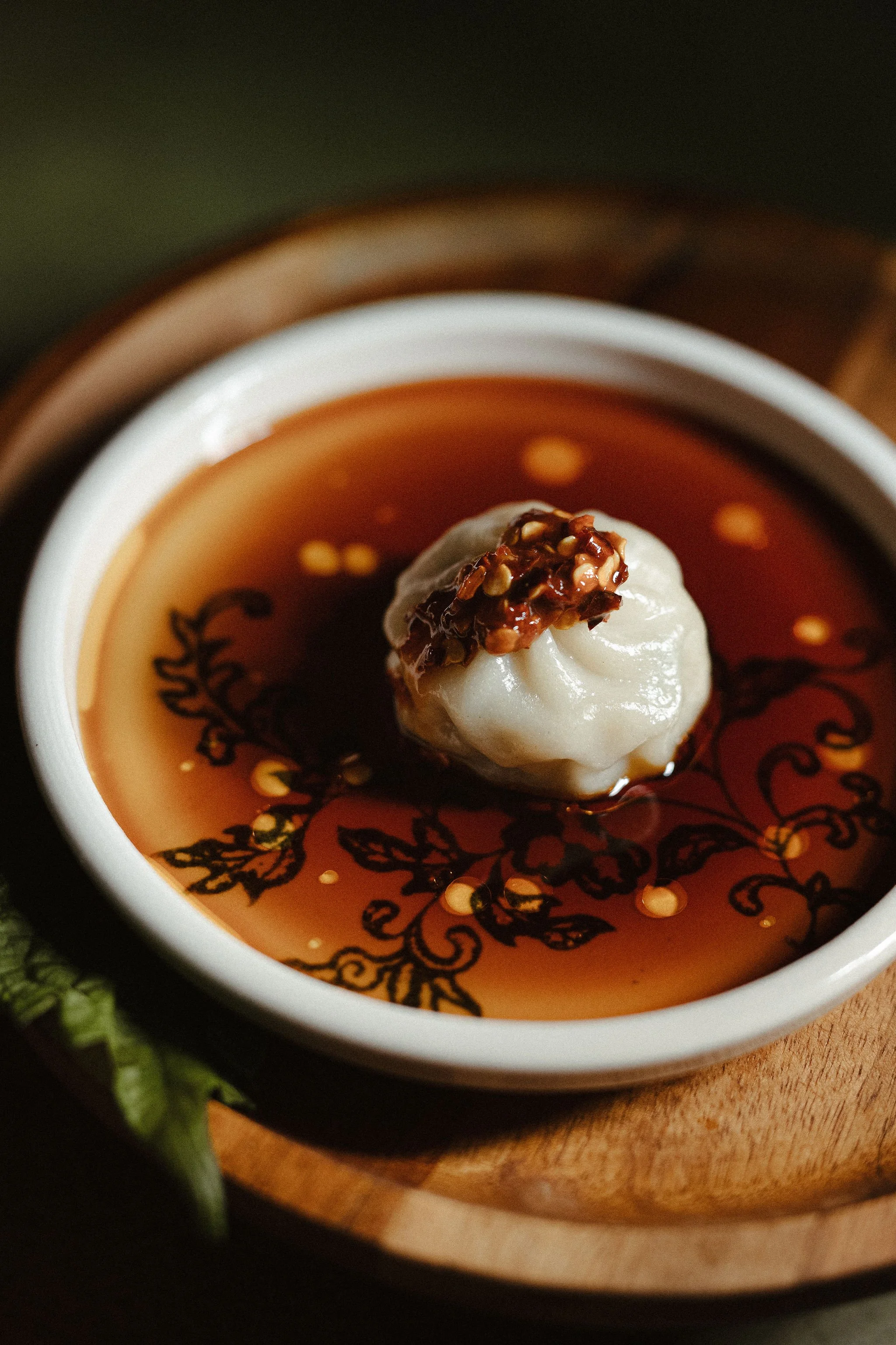 Close-up of a bowl of dark broth with a dumpling topped with chili flakes inside, with a decorative black pattern underneath.