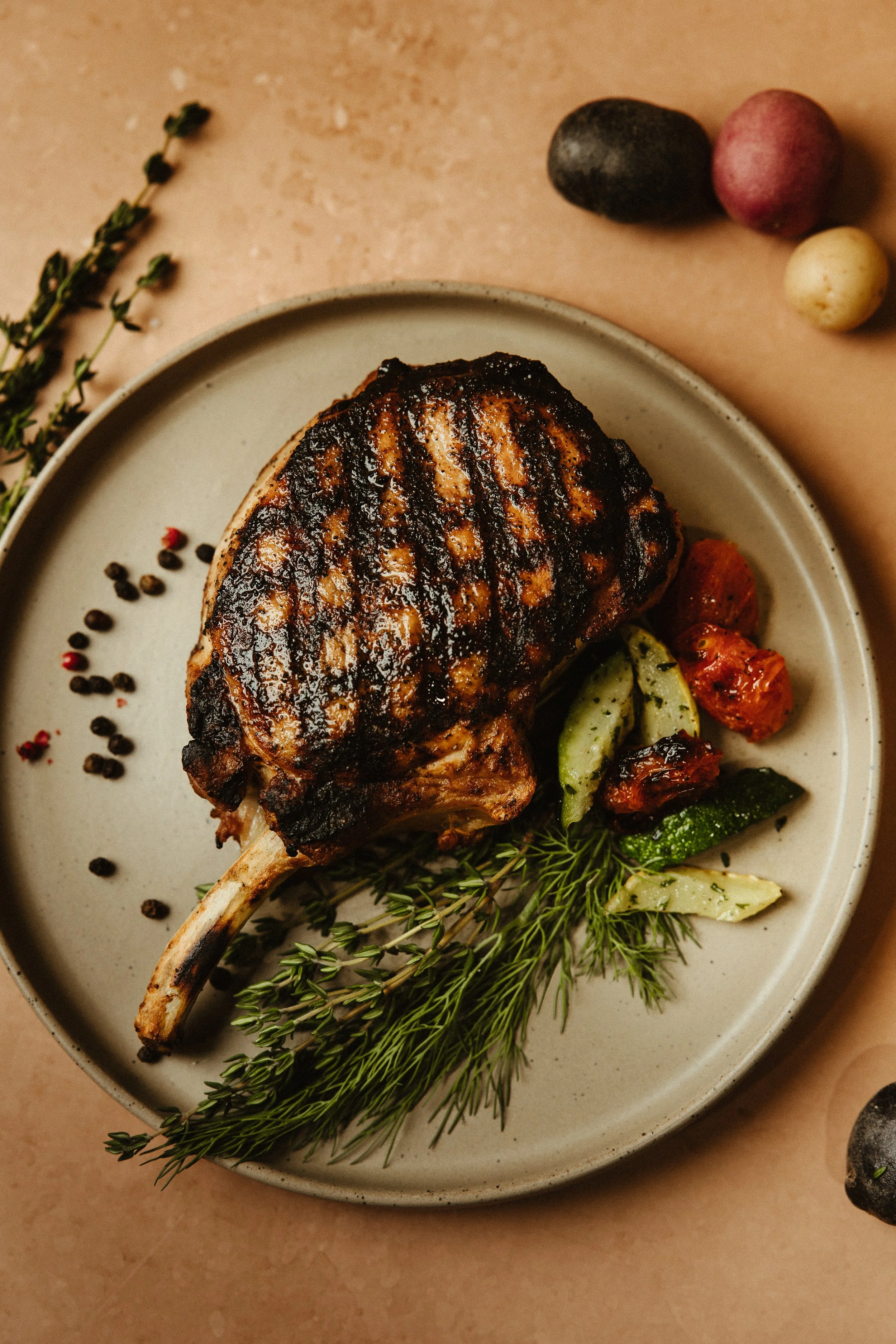 A grilled pork chop on a plate garnished with fresh herbs and vegetables, with black peppercorns scattered nearby and small potatoes and sprigs of herbs on the table.