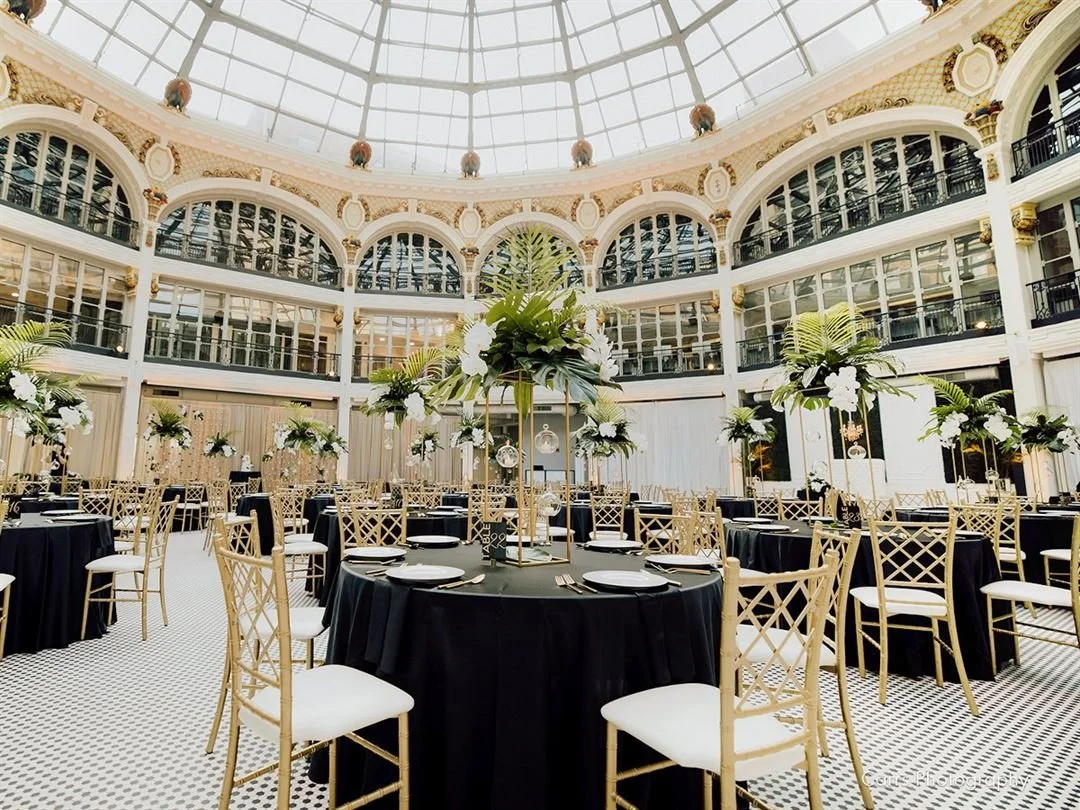 Elegant banquet hall with round tables covered in black cloth, decorated with tall floral centerpieces, set with white plates and silverware. The hall features a glass dome ceiling and a multi-level balcony with ornate architecture.