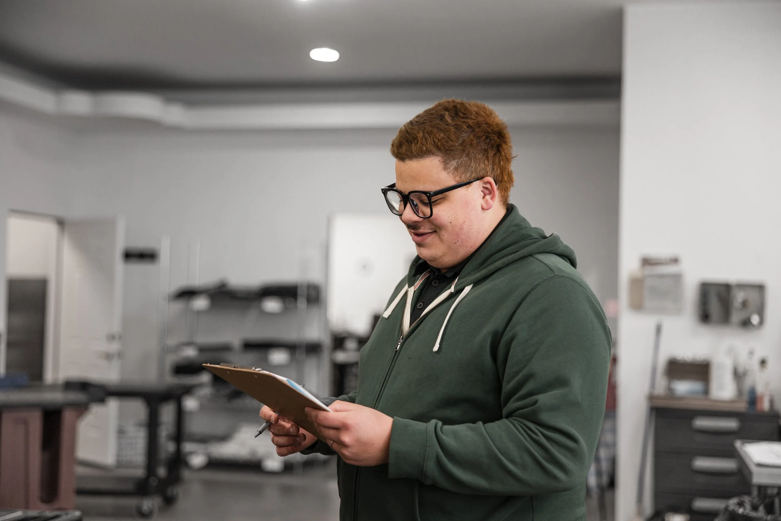 A young man with curly red hair and glasses, wearing a dark green hoodie, is holding a clipboard and a pen while standing in a kitchen, surrounded by appliances and counters.