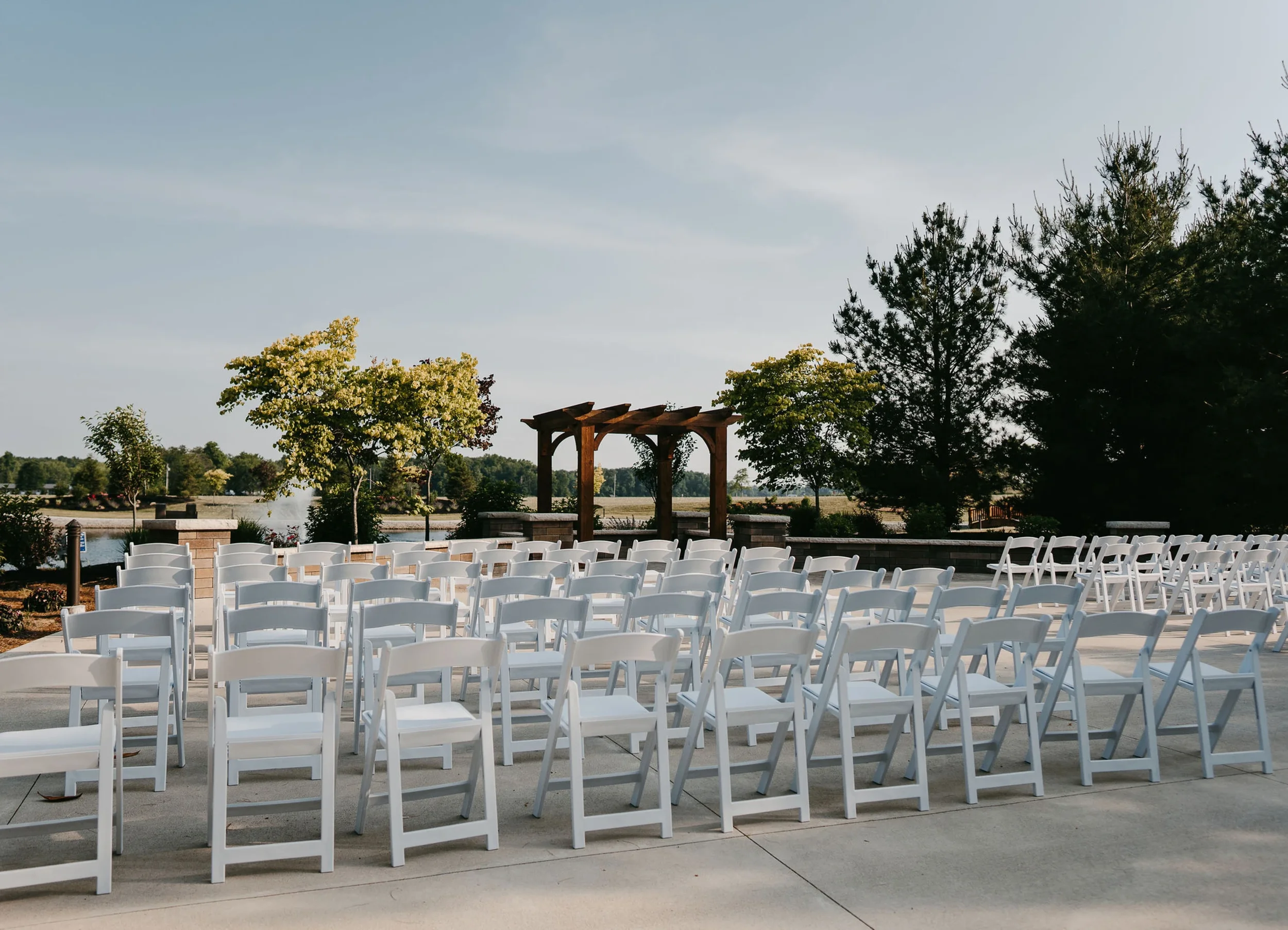 Outdoor wedding ceremony setup with white chairs arranged in rows on concrete, a wooden gazebo, trees, a lake, and a slightly cloudy sky.