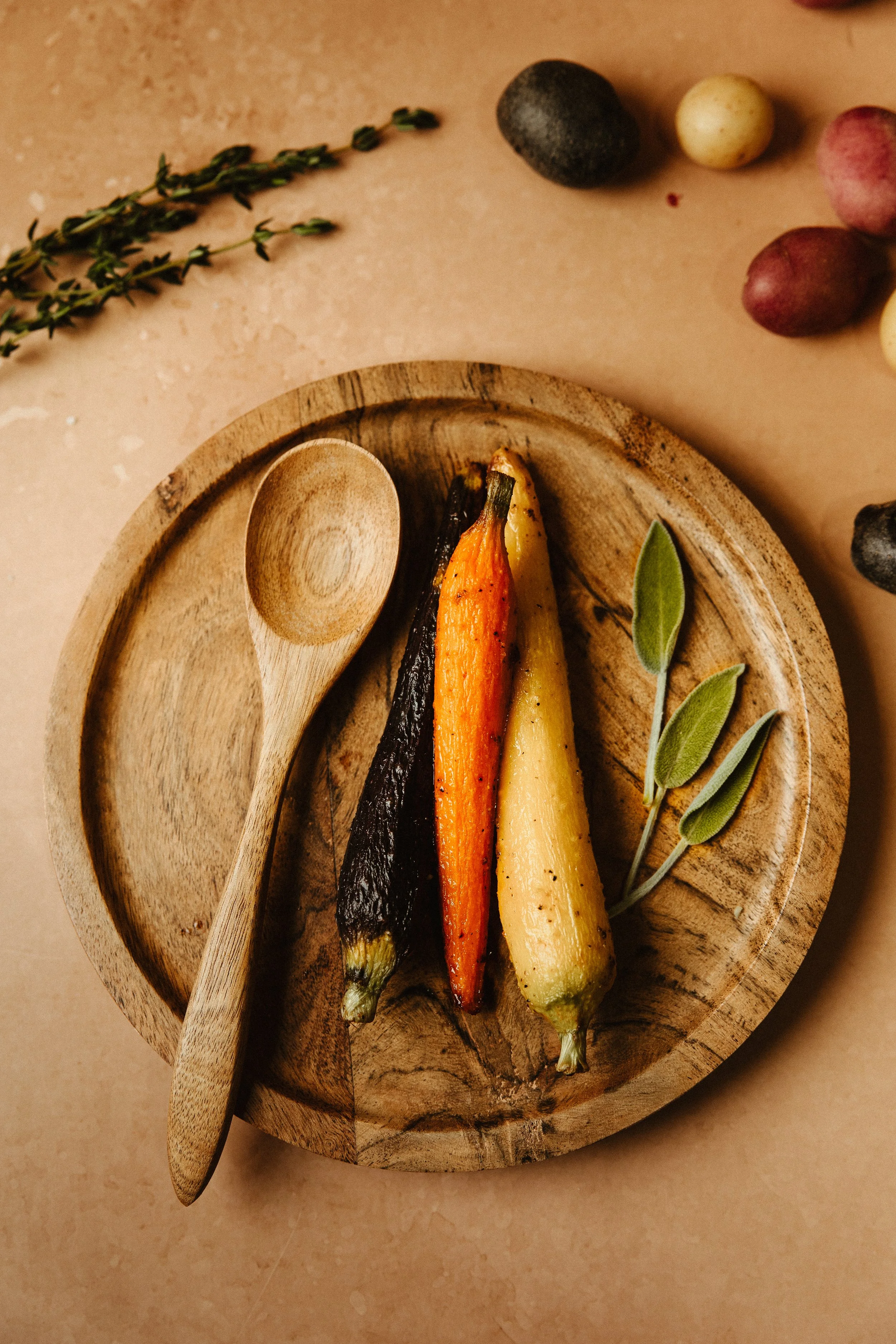 Roasted multicolored carrots with fresh sage leaves on a round wooden plate, with a wooden spoon resting on the edge of the plate, and fresh herbs and vegetables in the background.